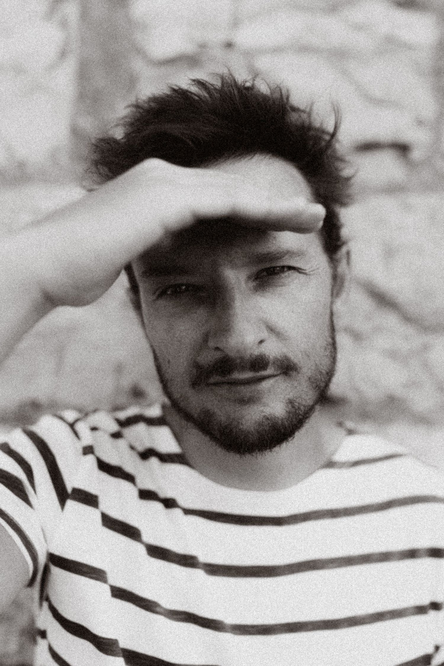 Black and white portrait of a young bearded man in a striped shirt shielding his eyes with his hand outdoors.