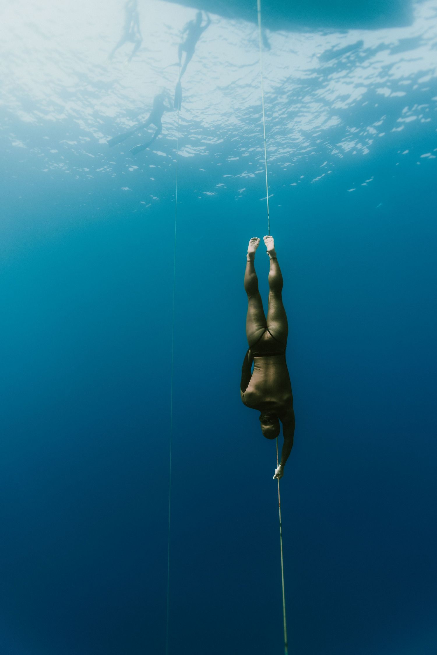 A freediver descends into deep blue ocean waters while holding onto a guide rope against a sunlit underwater backdrop.