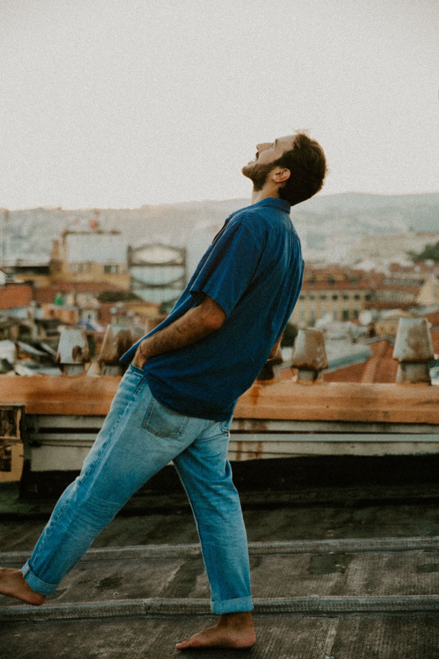 Person in blue shirt and jeans dancing on a rooftop with cityscape in background.
