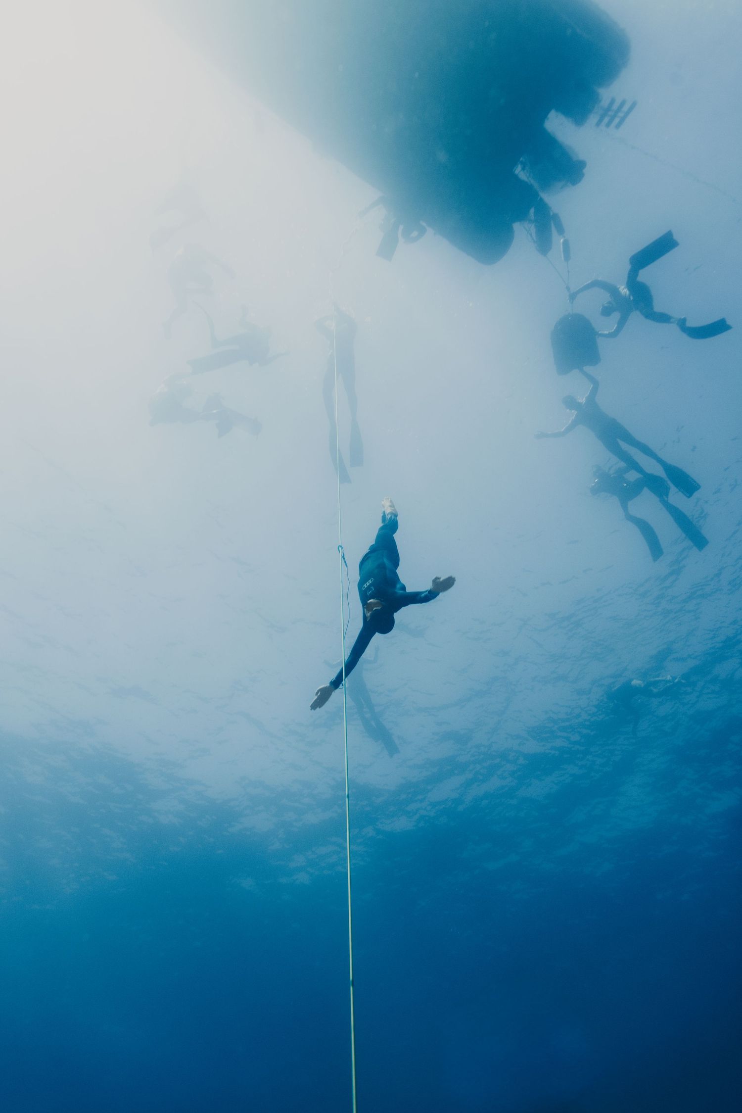 Underwater view of freedivers descending on a rope in clear blue ocean water with a boat visible at the surface.
