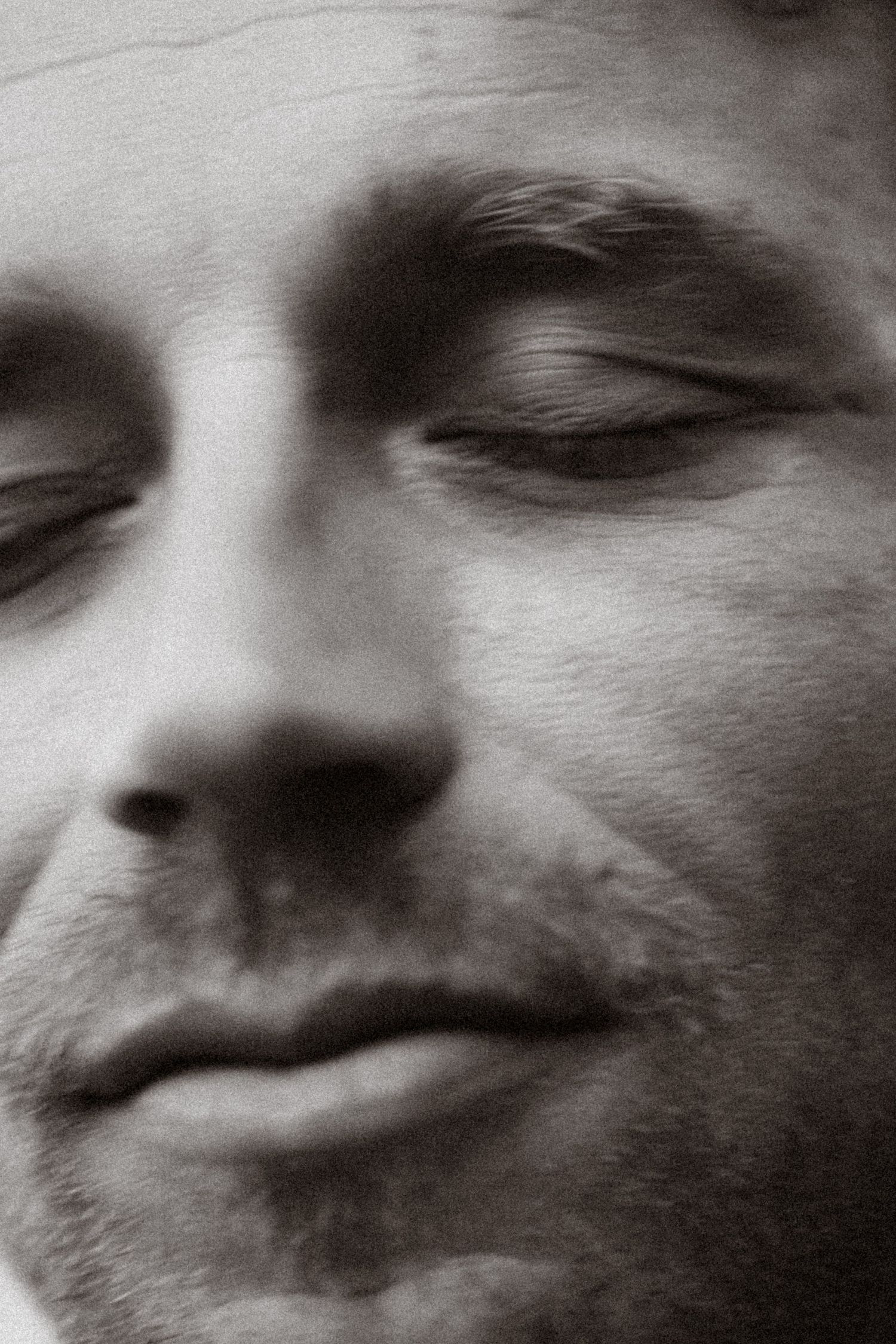 Close-up black and white portrait of a man with closed eyes and slight smile, showing textured skin detail.