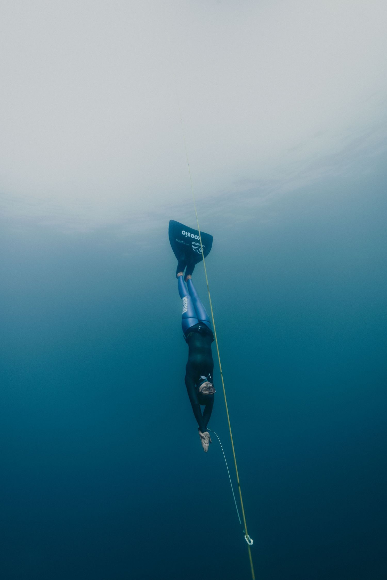 Freediver in wetsuit and fins descends along a rope into deep blue ocean water.