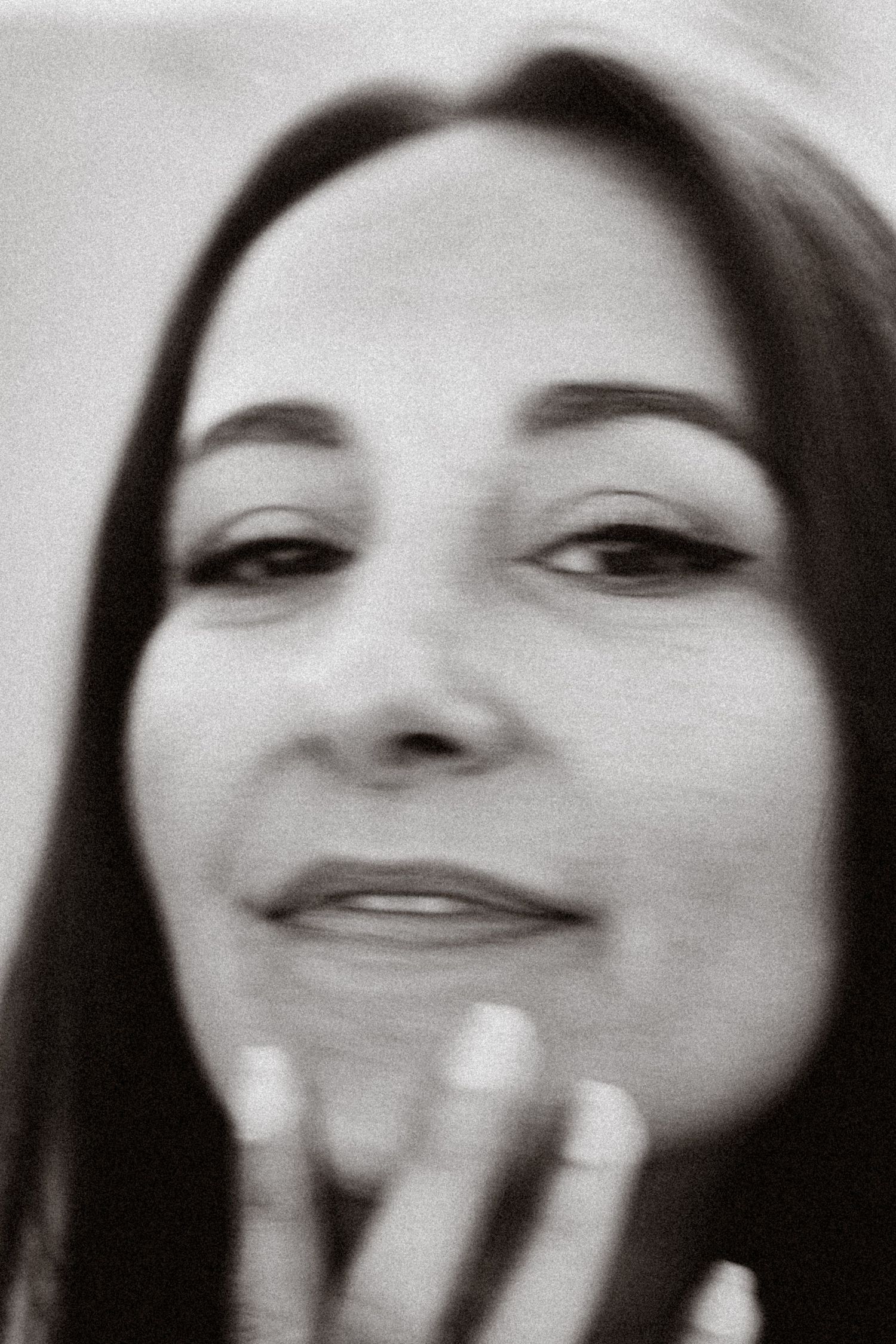 Black and white close-up portrait of a woman with long dark hair, smiling softly with hand near face.