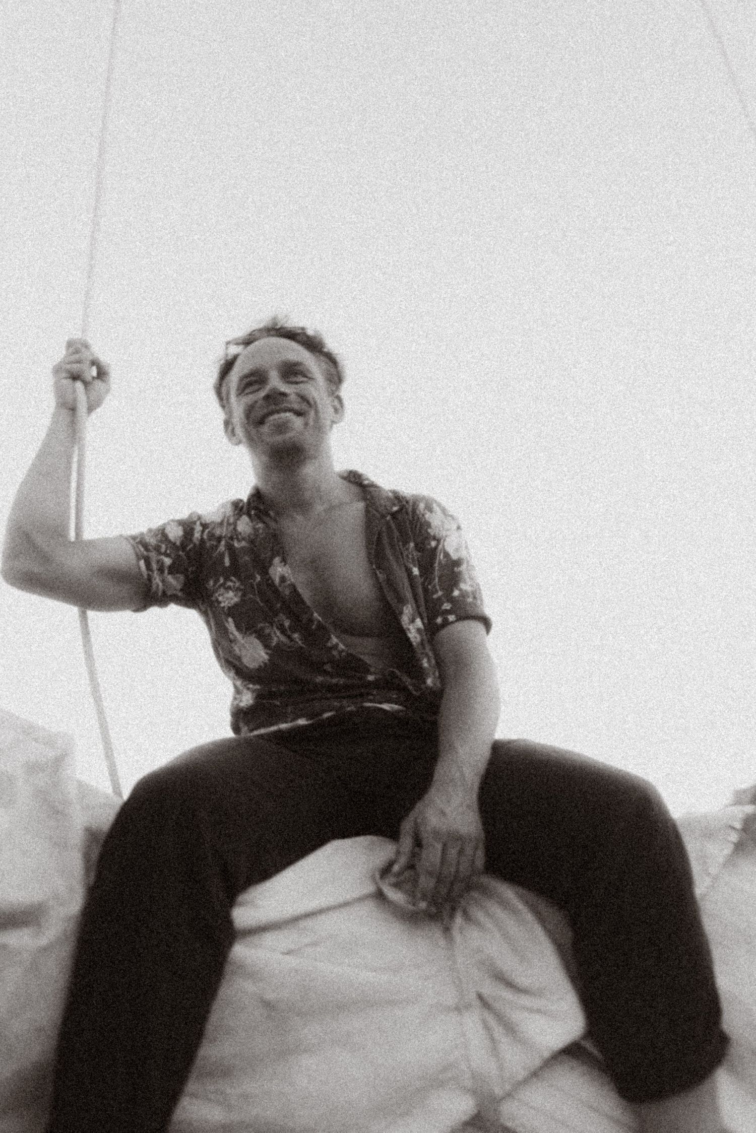 Smiling man in floral shirt sits on sailboat rigging, black and white vintage photo taken from below.
