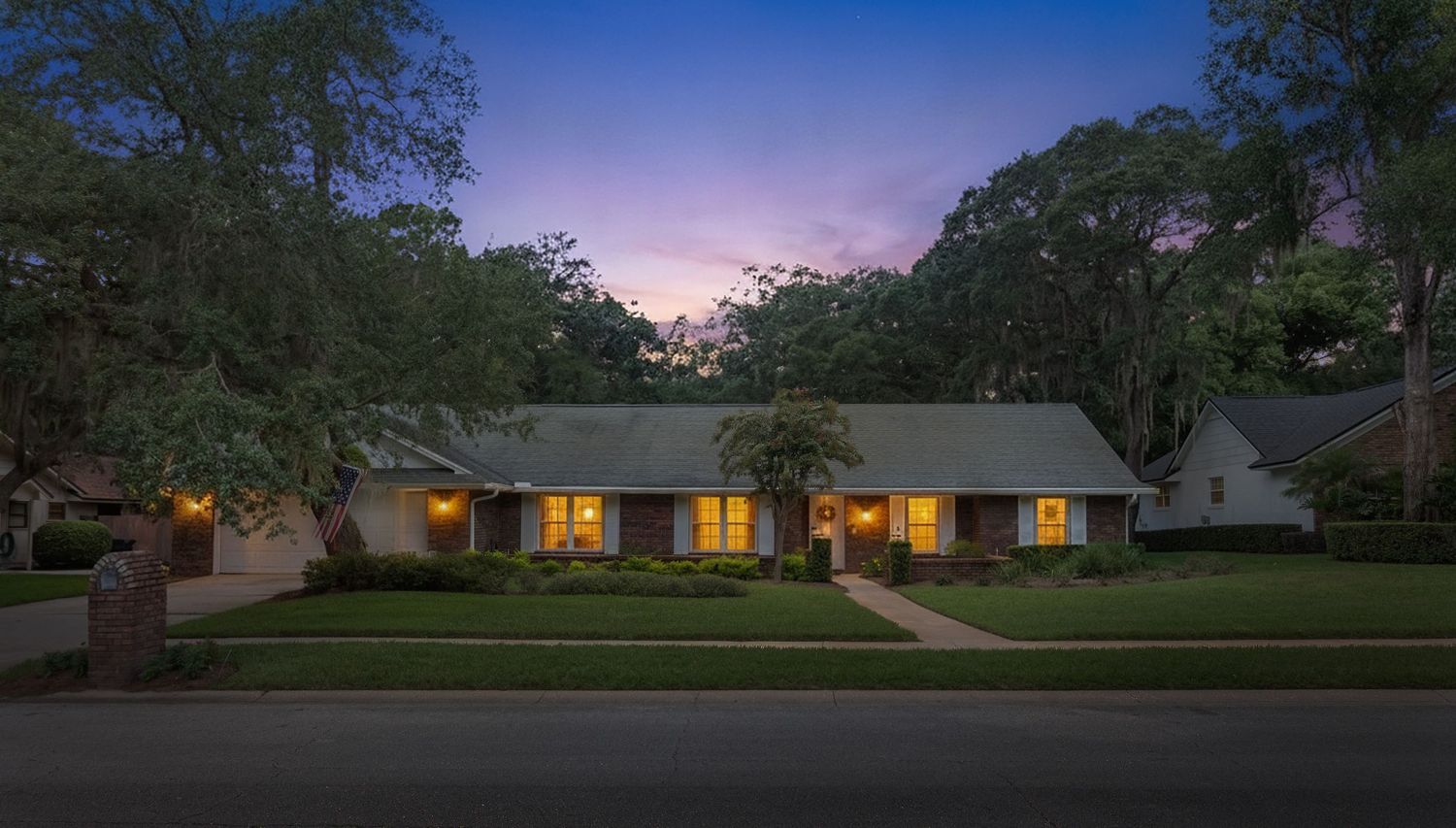 Twilight real estate photography of a single-story home with glowing windows, a landscaped front lawn, and a purple sunset sky.