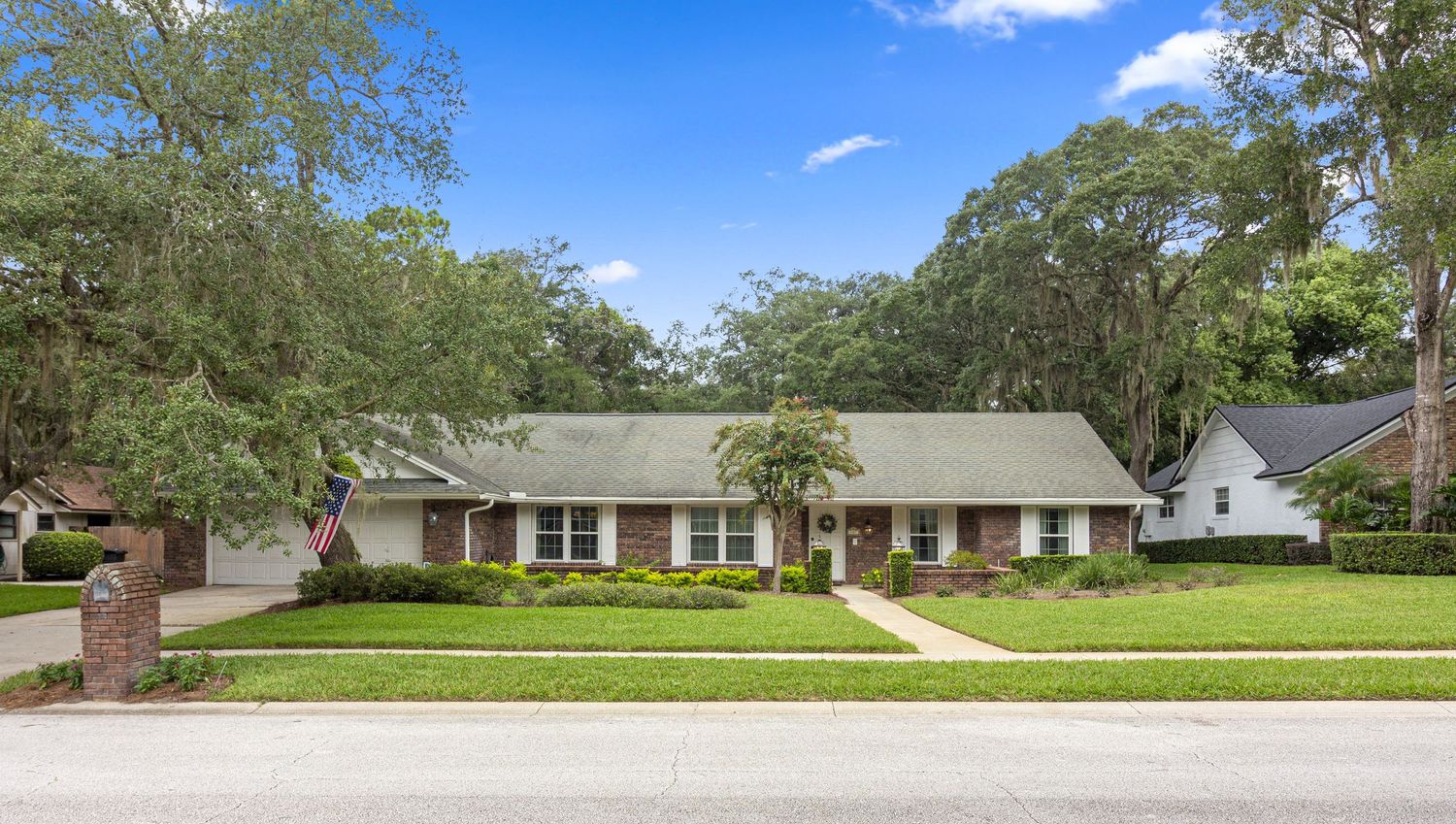 Daytime exterior of a single-story brick ranch home with a manicured lawn, mature trees, and a clear blue sky.