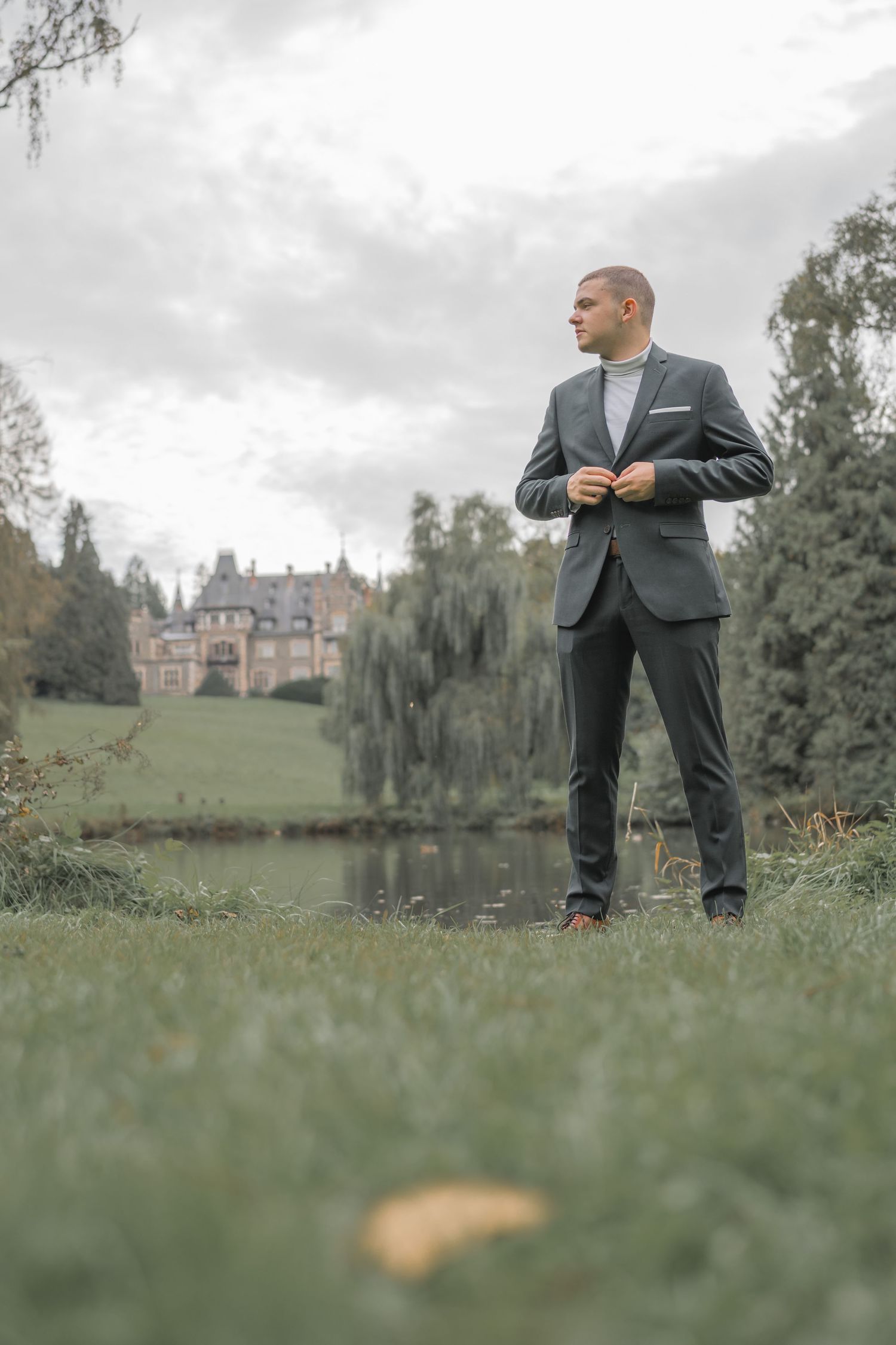 Person in a suit standing on grass with a grand castle and lake in the background.