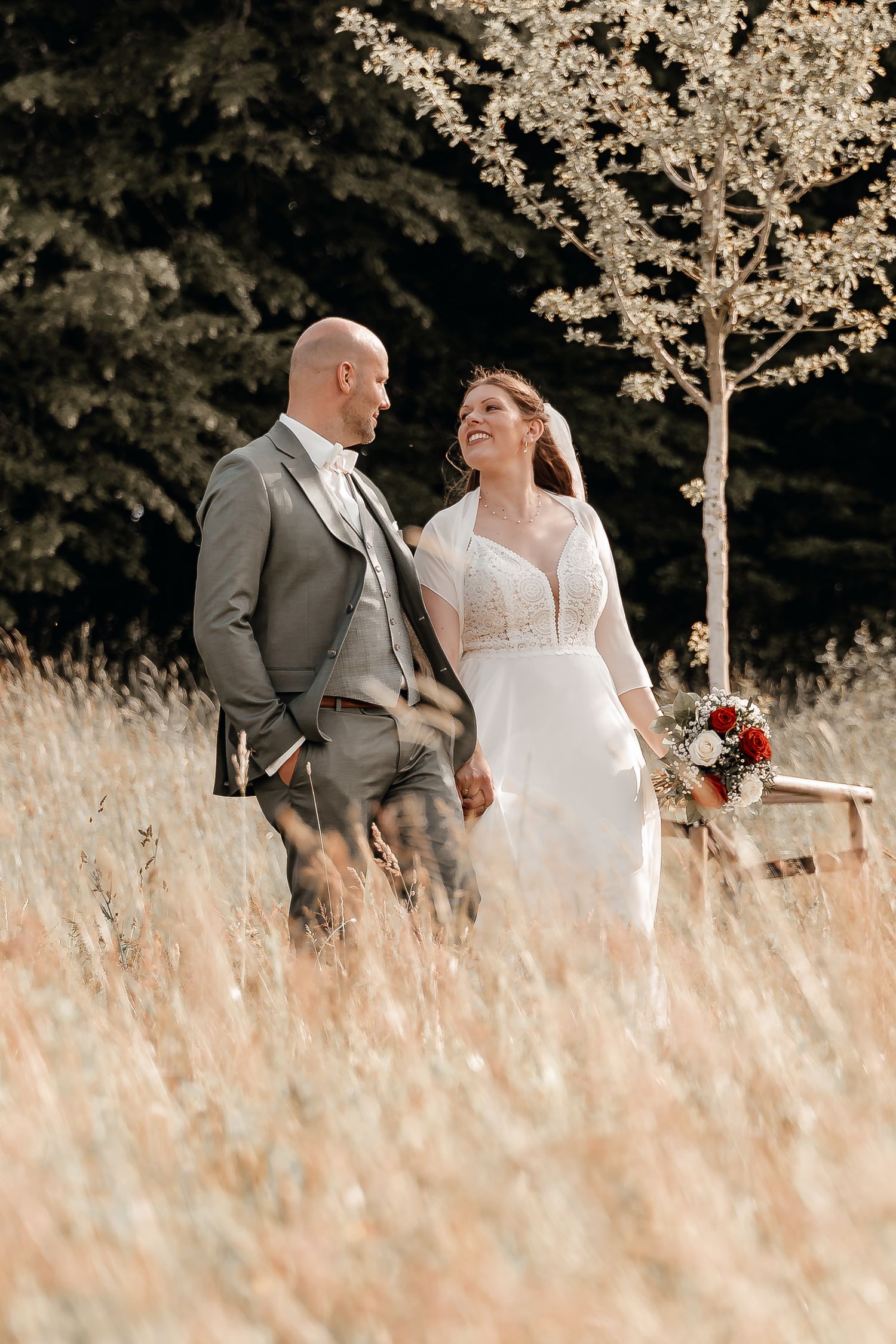 A smiling bride in a white gown and groom in a gray suit walk through a golden meadow on their wedding day.