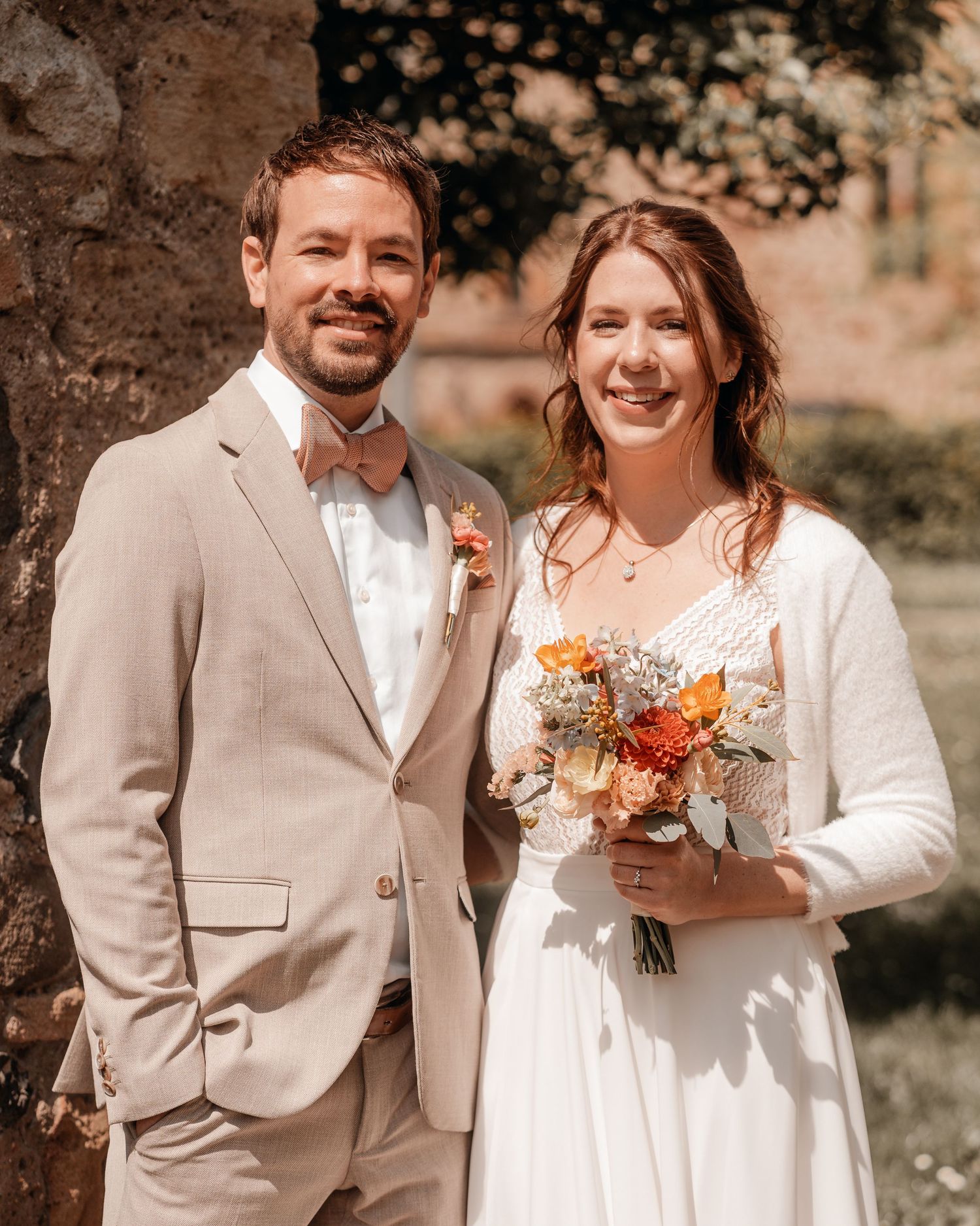 Couple in coordinated white and beige wedding attire posing with a colorful bouquet.