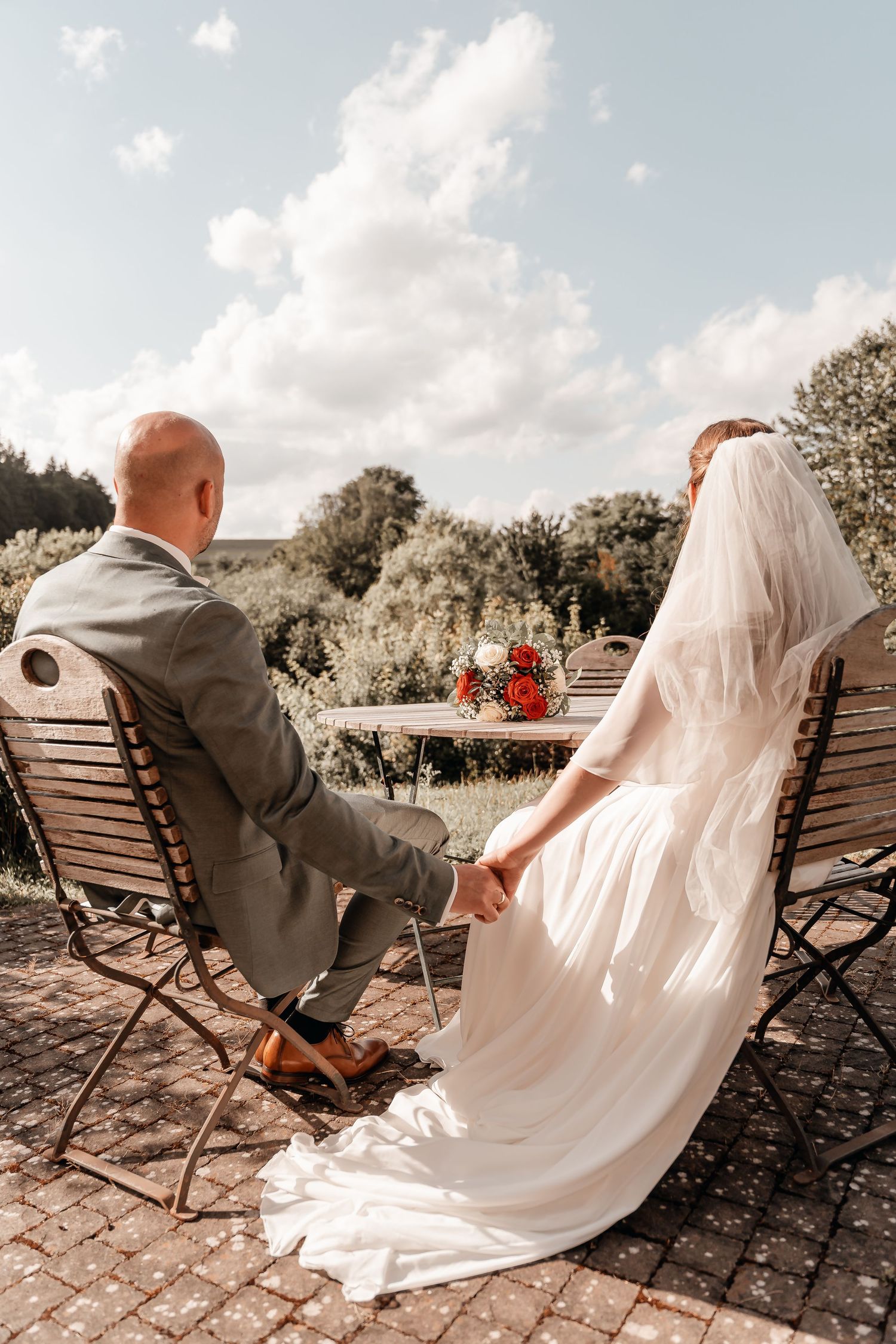 Bride and groom holding hands at outdoor table, bride in white gown with veil, groom in gray suit, on sunny day.