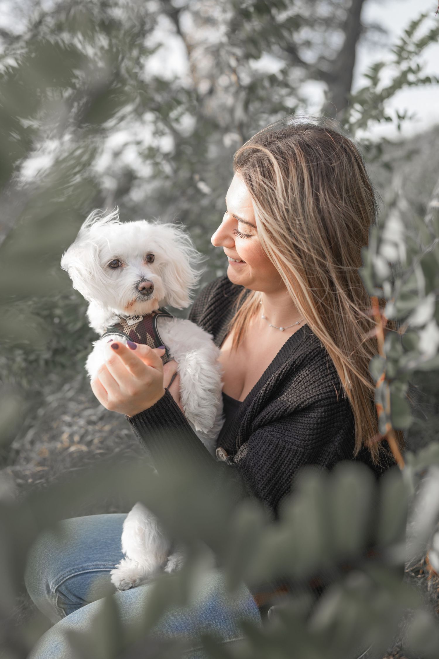Woman in black sweater holding a small white fluffy dog among tree branches.