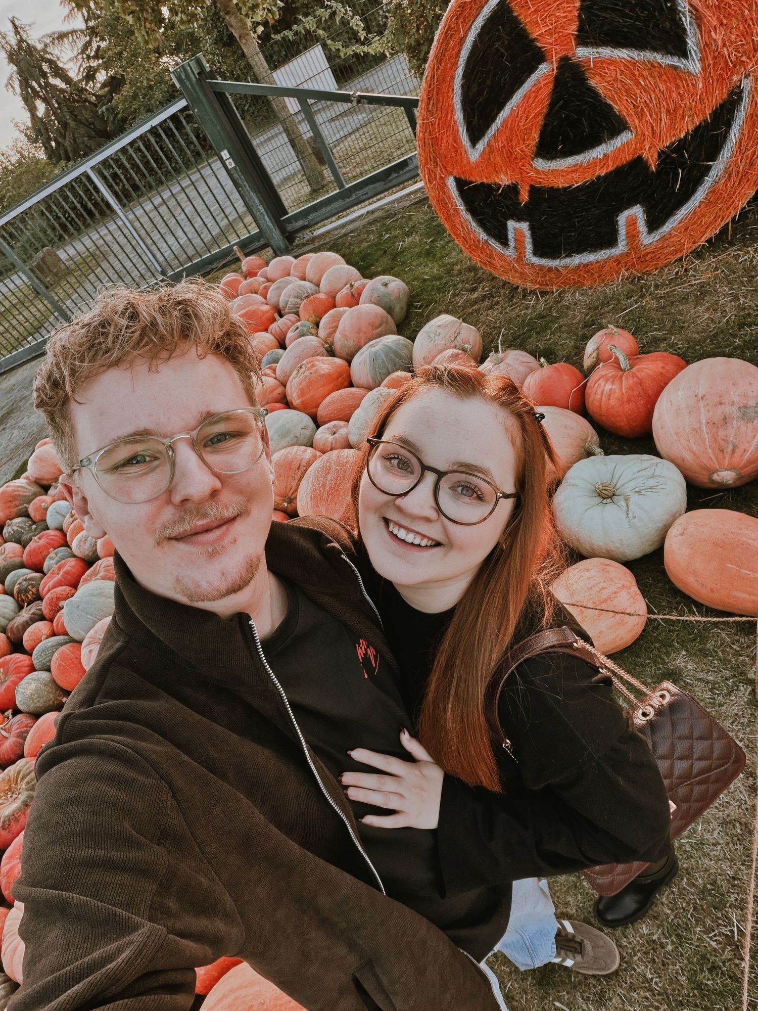 Couple smiling for selfie in front of colorful pumpkin patch display with jack-o-lantern hay bale decoration.