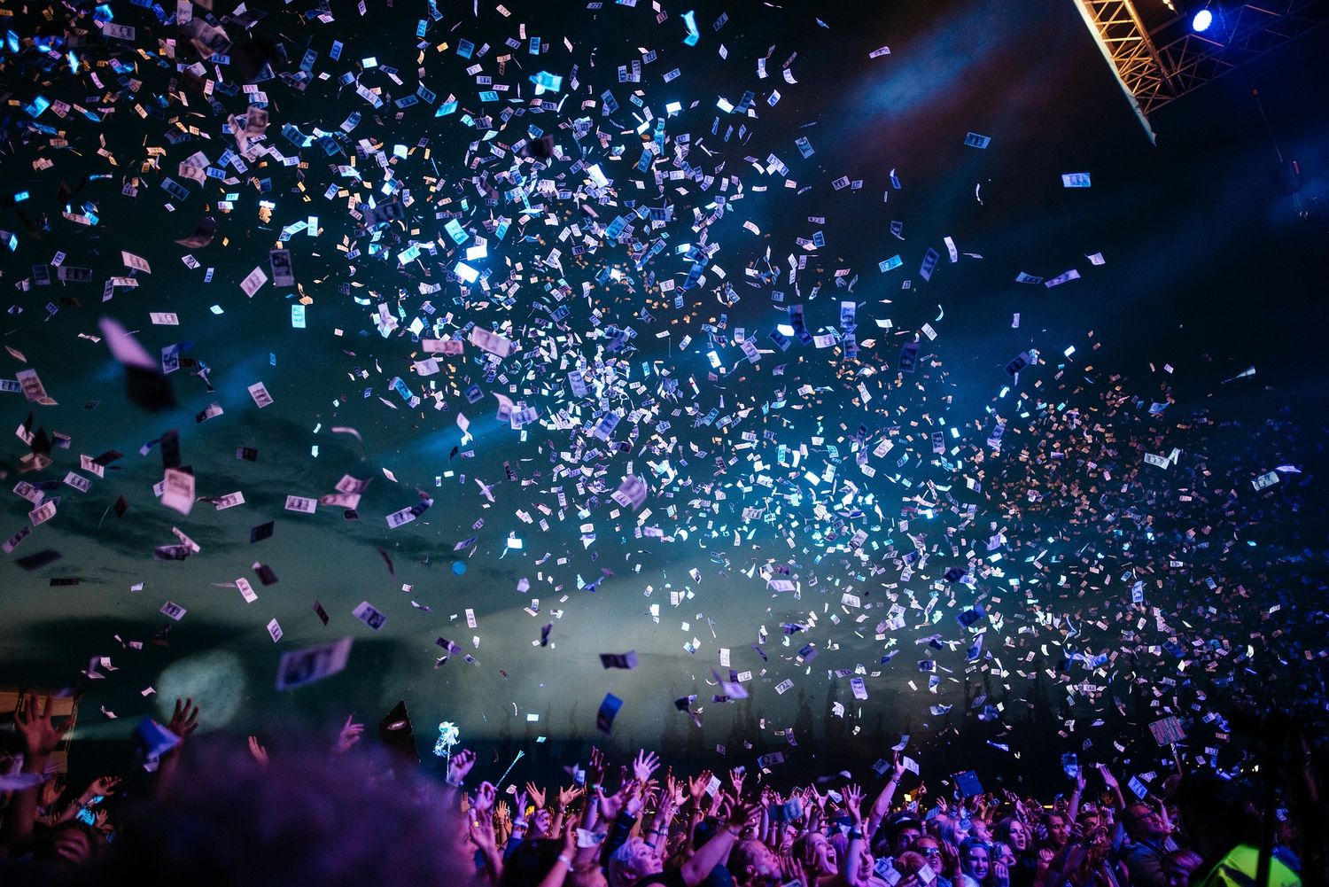 Concert crowd with raised hands under falling confetti and blue lighting during a live music performance.