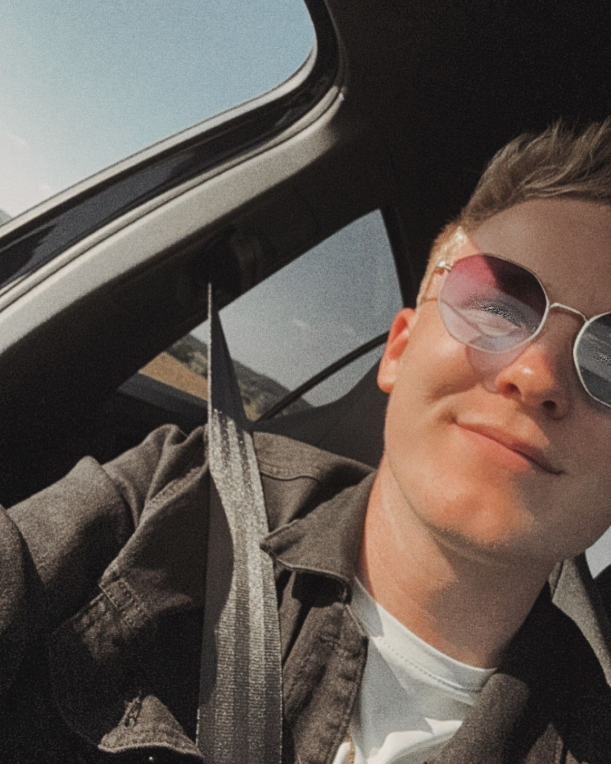Person relaxing in car wearing sunglasses with natural light streaming through window.