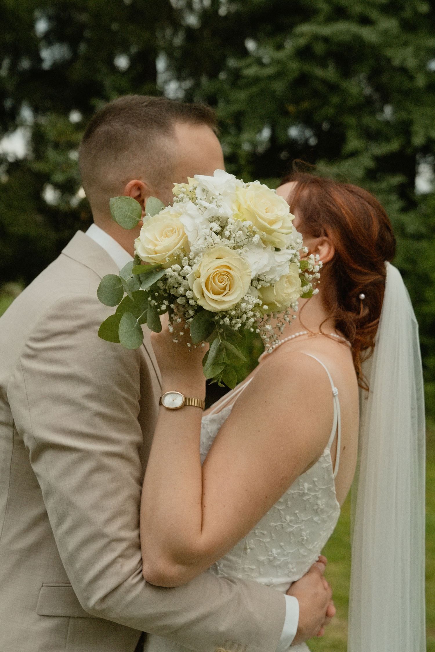 Bride and groom sharing a kiss hidden behind a white rose bouquet outdoors on their wedding day.