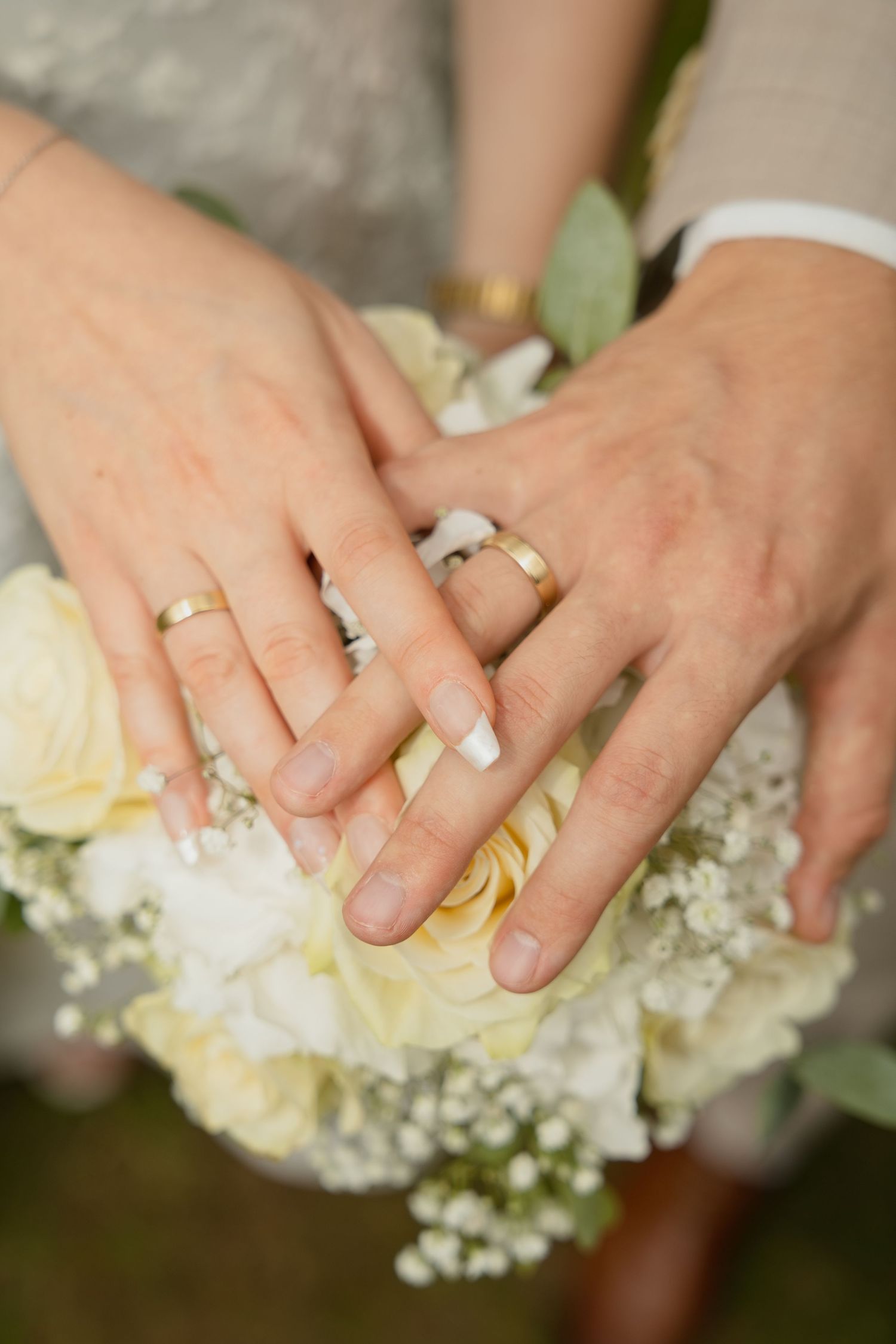 Newlywed couple displaying gold wedding rings on hands resting over a white rose and baby's breath bridal bouquet.