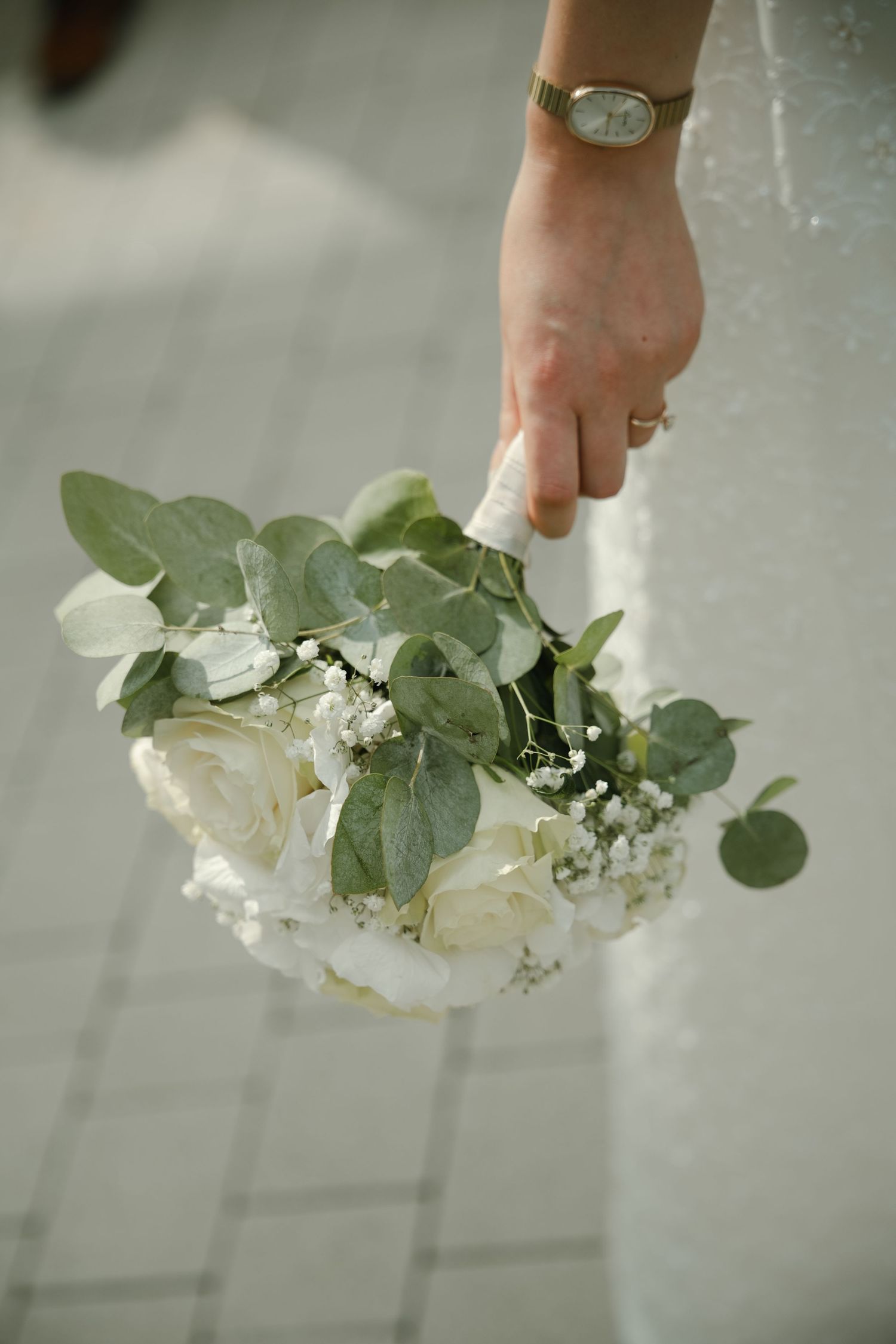 Bride holding a white rose and eucalyptus wedding bouquet with baby's breath, wearing a gold watch and ring.