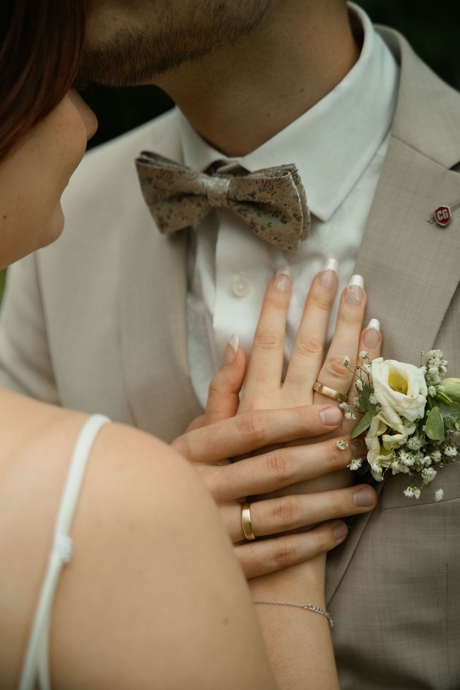 Bride and groom close-up showing wedding rings, french manicure, boutonniere, and beige suit with bow tie.