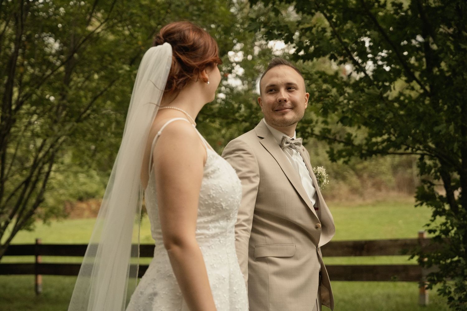 Bride in white gown and veil stands with groom in tan suit outdoors at a wedding ceremony near a wooden fence.