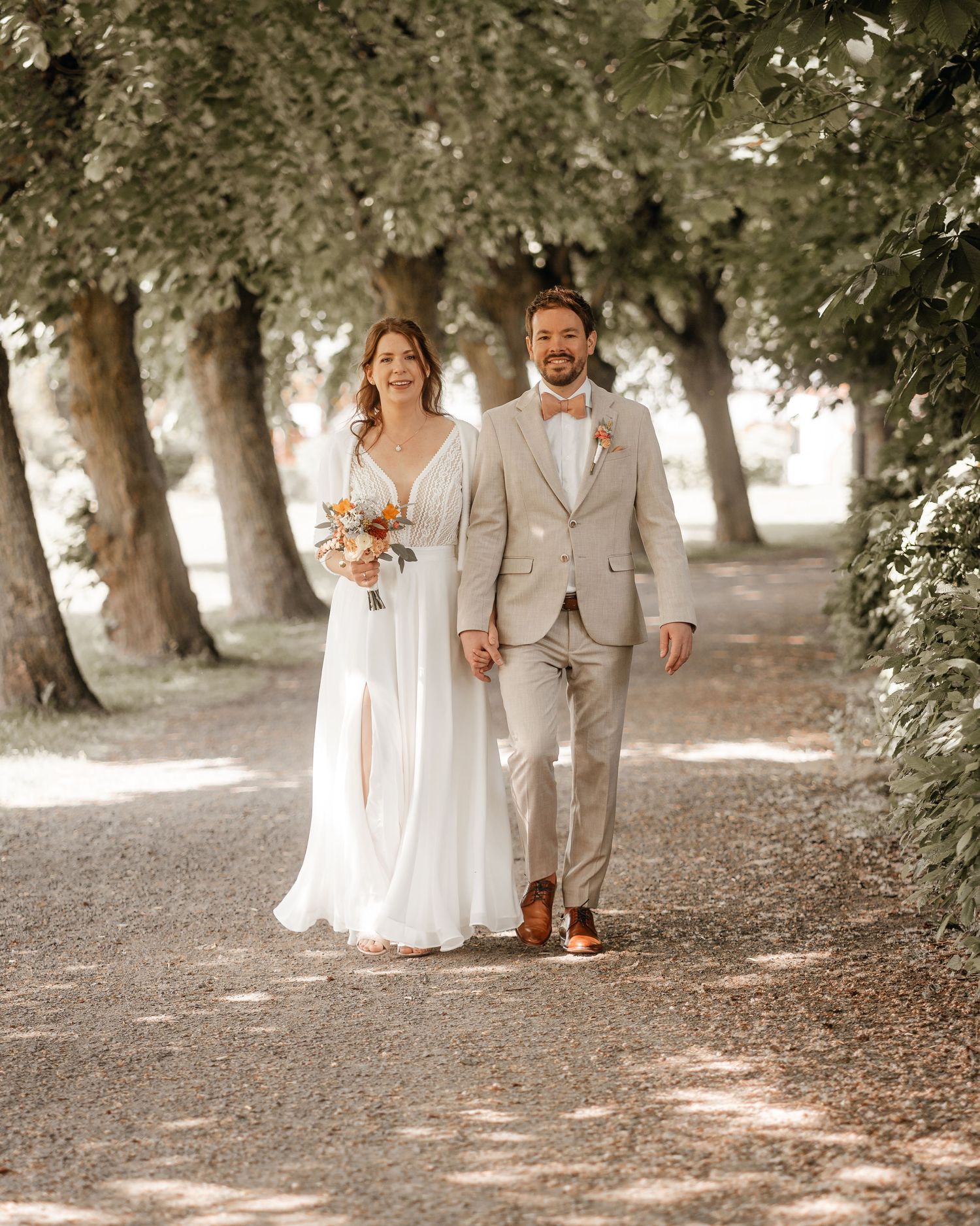 Newlyweds strolling through a scenic park path surrounded by mature trees.