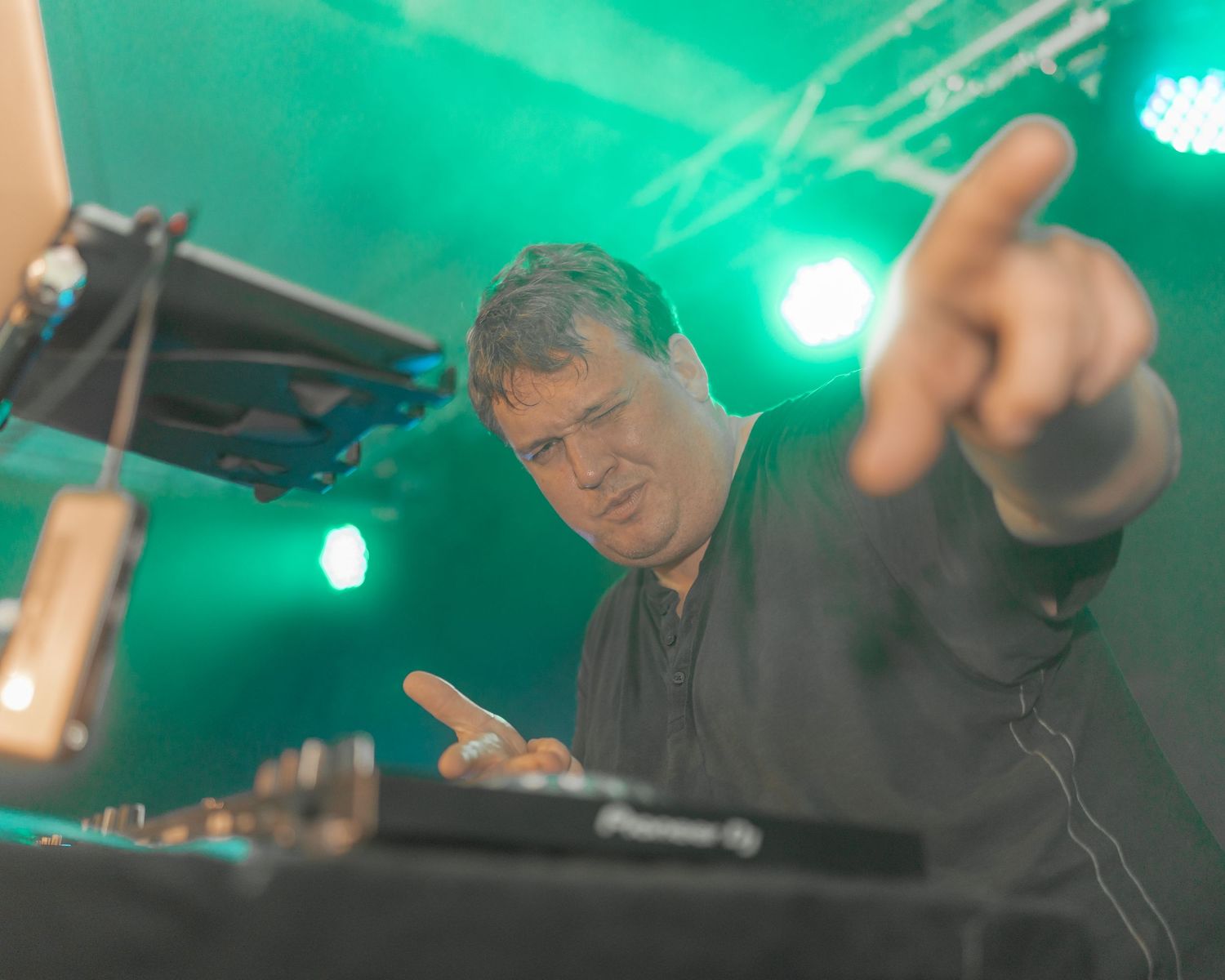 DJ performing at mixing desk under green stage lighting with animated gestures.