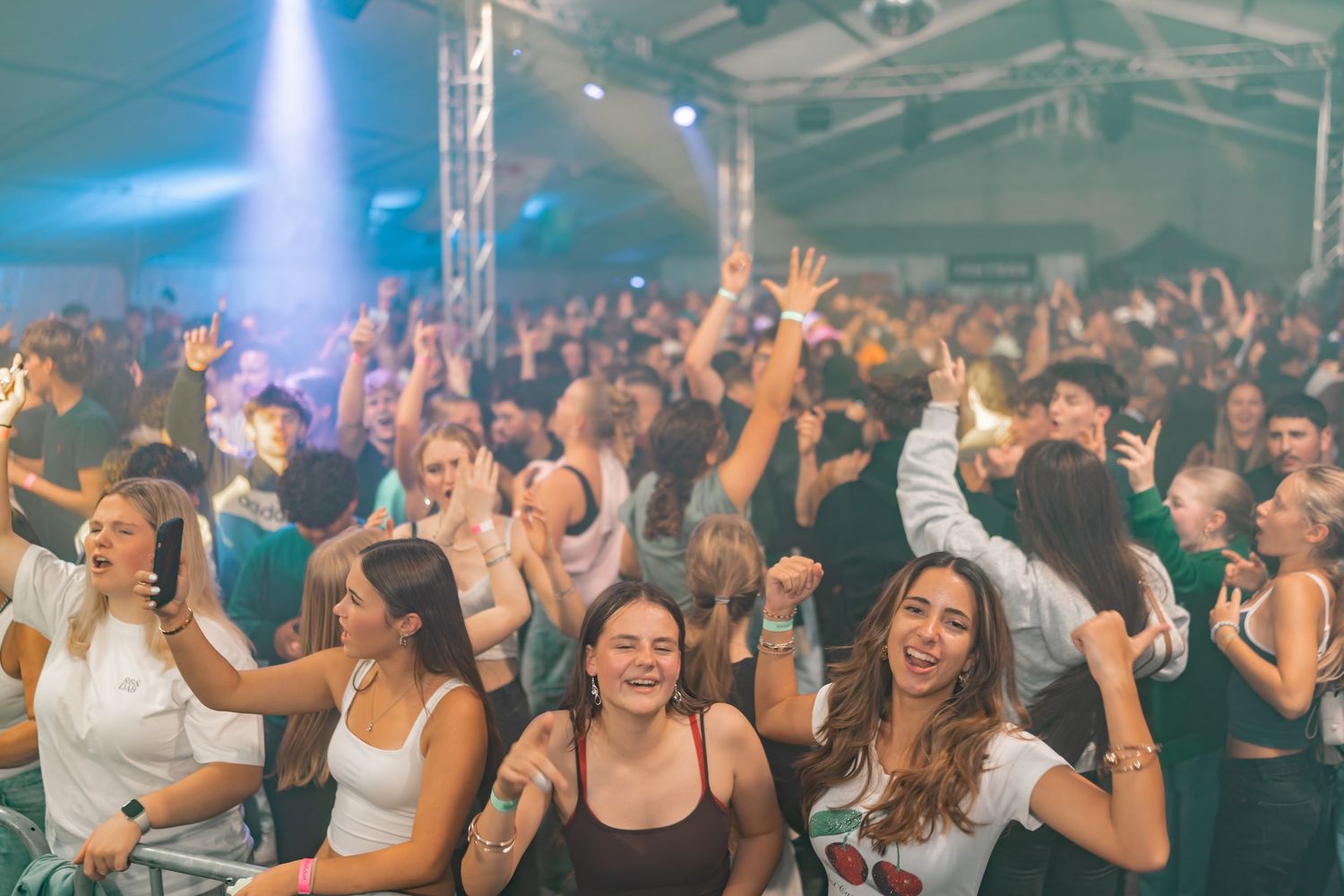 Energetic crowd dancing with raised hands at a lively indoor concert venue under blue lighting.