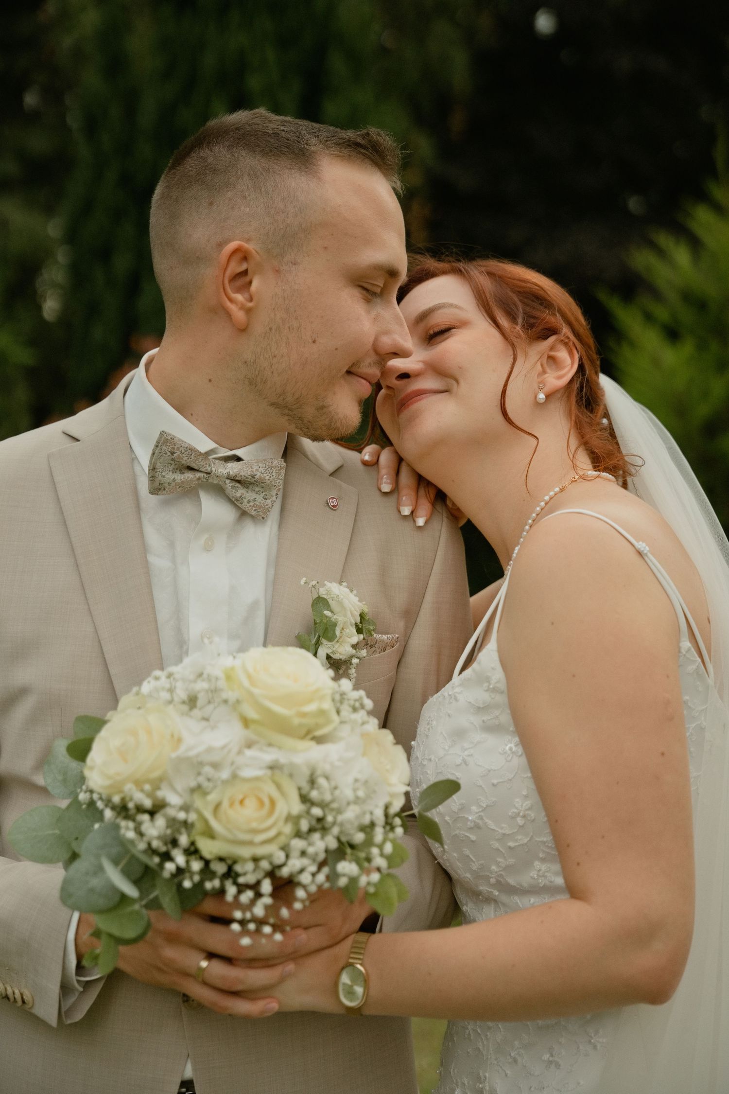 A smiling bride with red hair leans into her groom in a tan suit, holding a white rose bouquet outdoors.