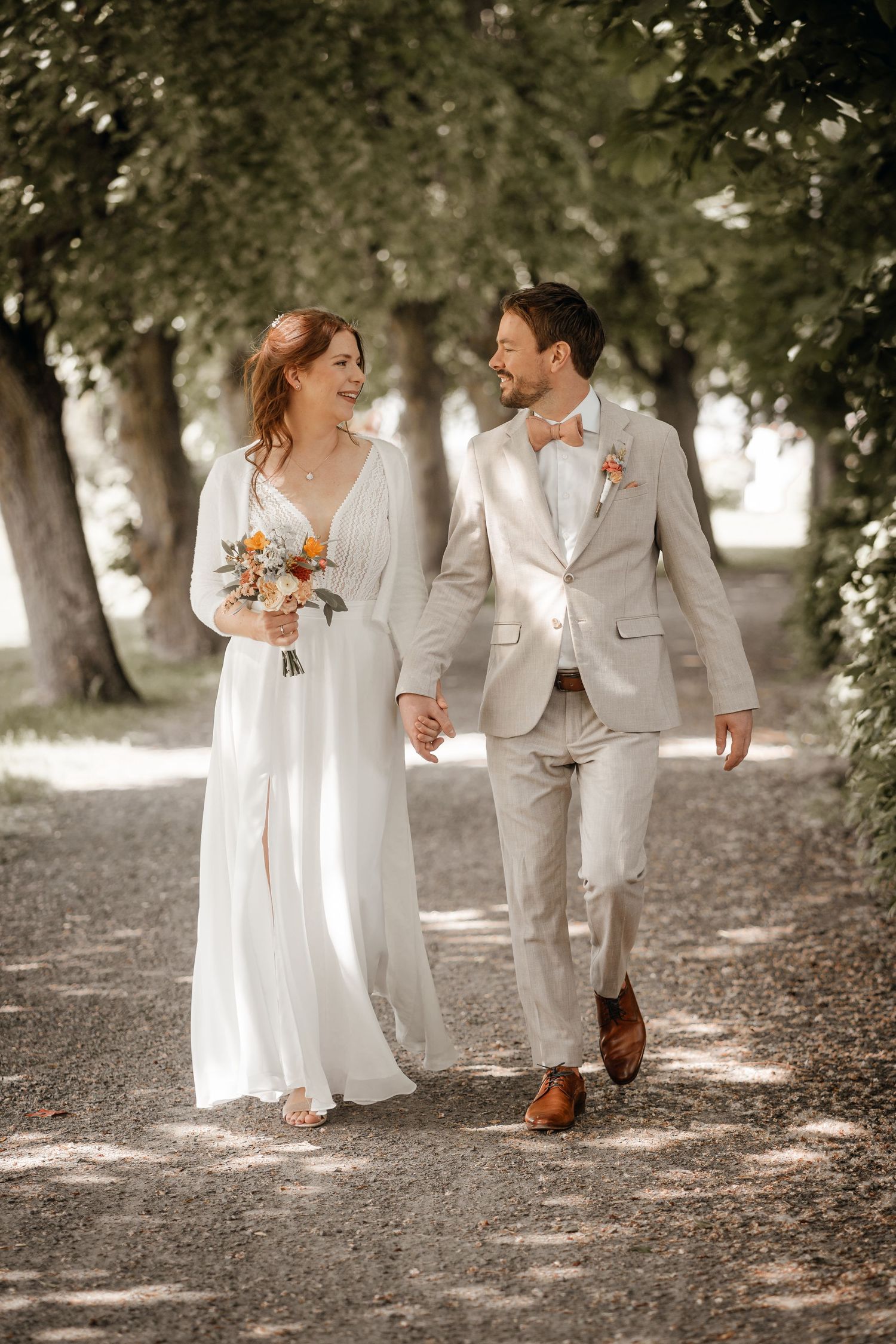Wedding couple in white and beige outfits strolling through a serene park setting.
