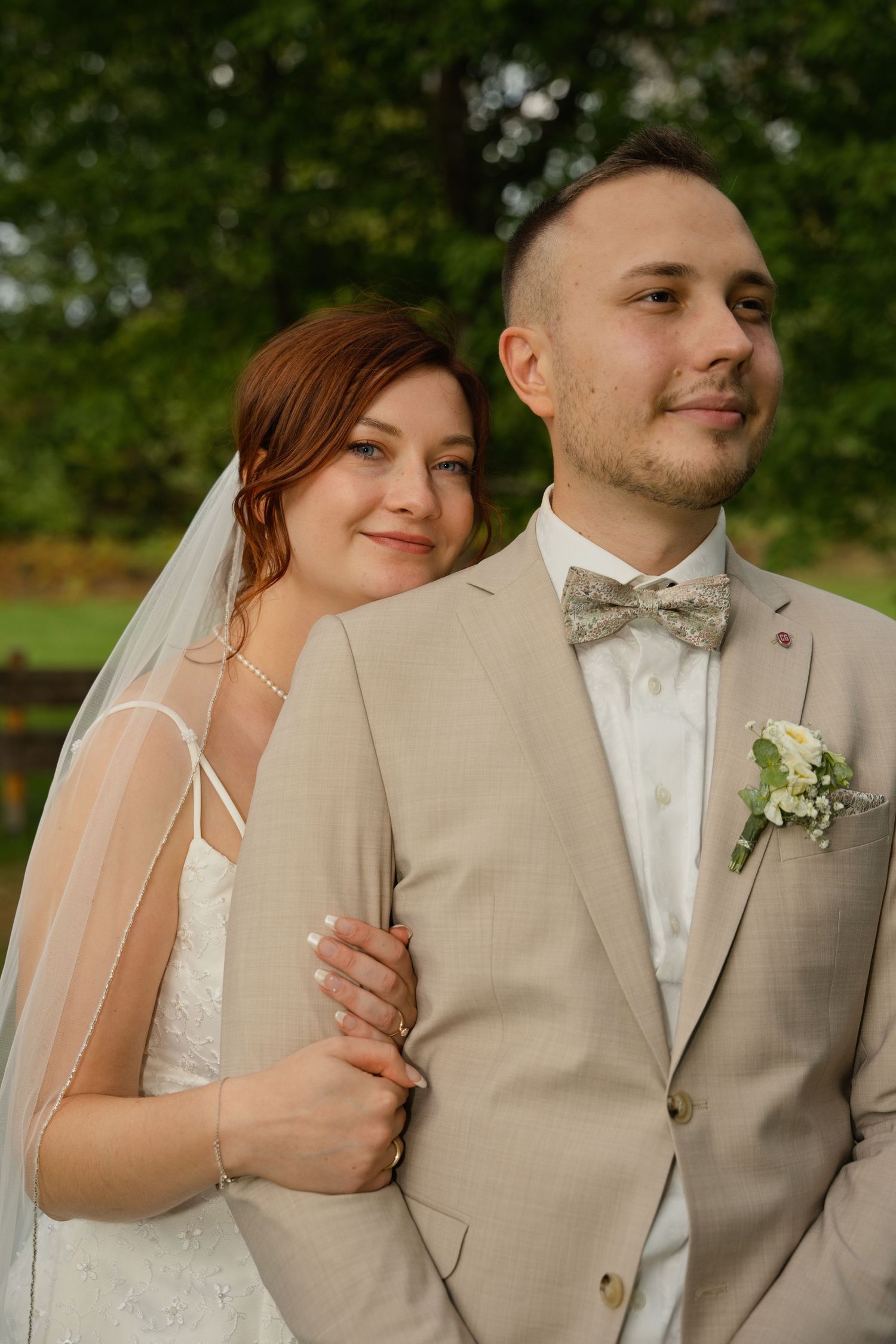 Bride in white dress and veil embraces groom in beige suit with bow tie and boutonniere outdoors.