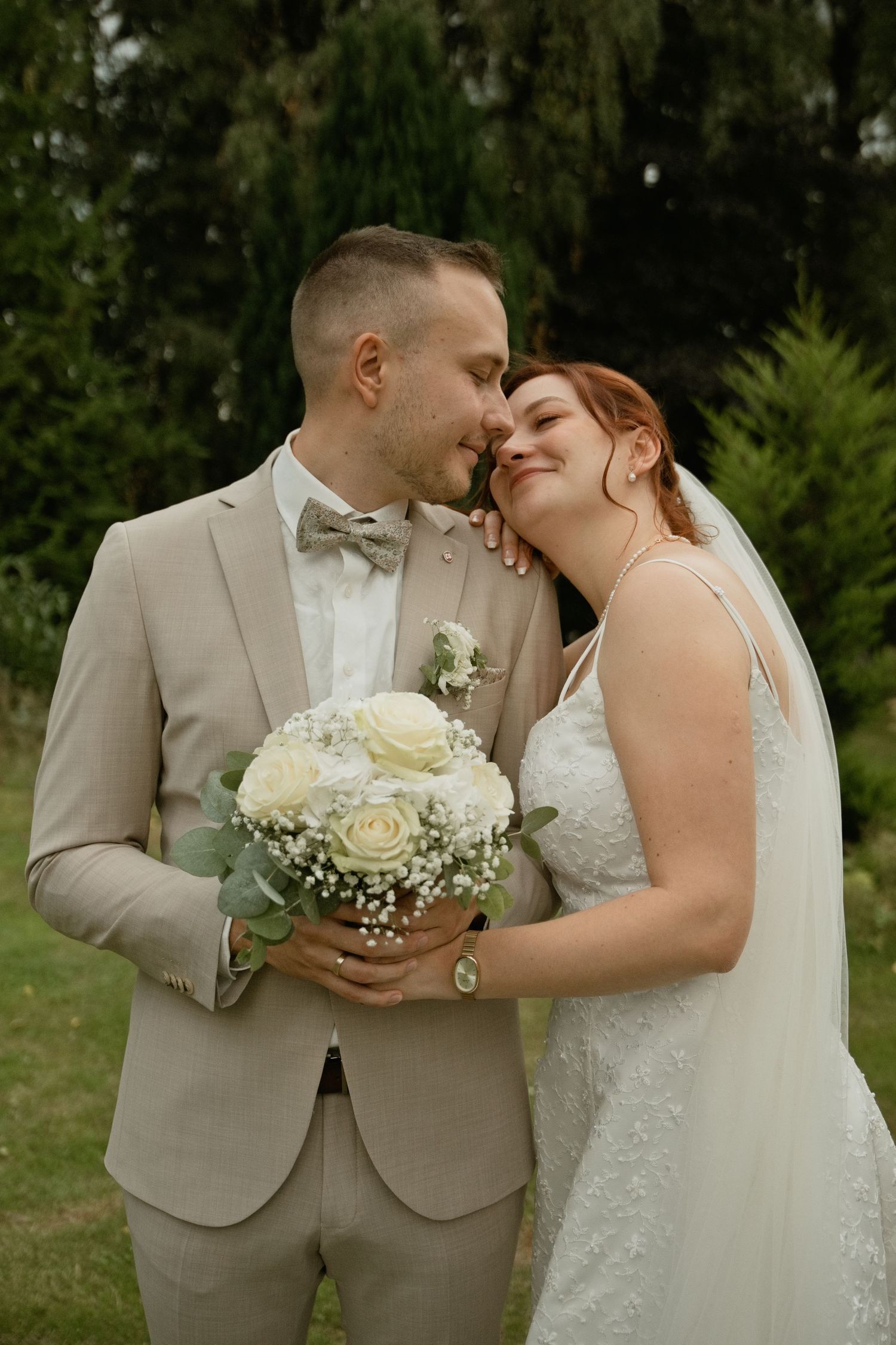 A bride and groom share a tender moment outdoors, holding white roses bouquet, groom in beige suit, bride in lace gown.