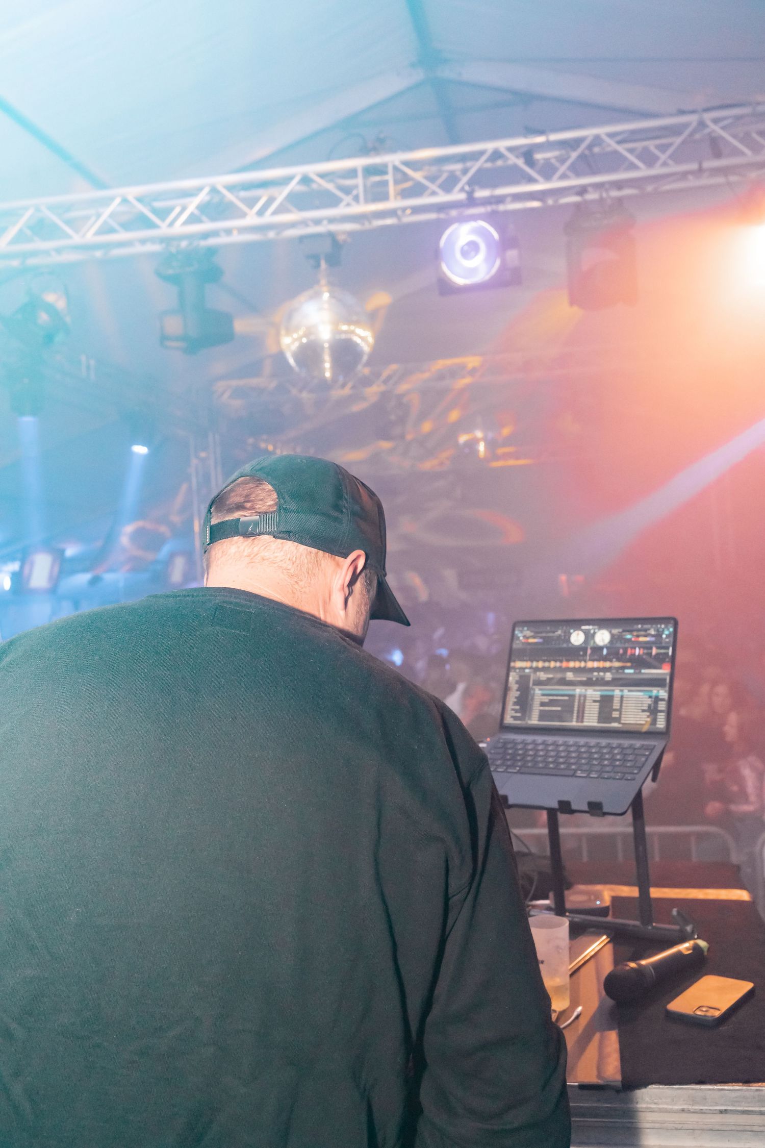 DJ performing at a concert venue seen from behind with equipment and stage lighting.