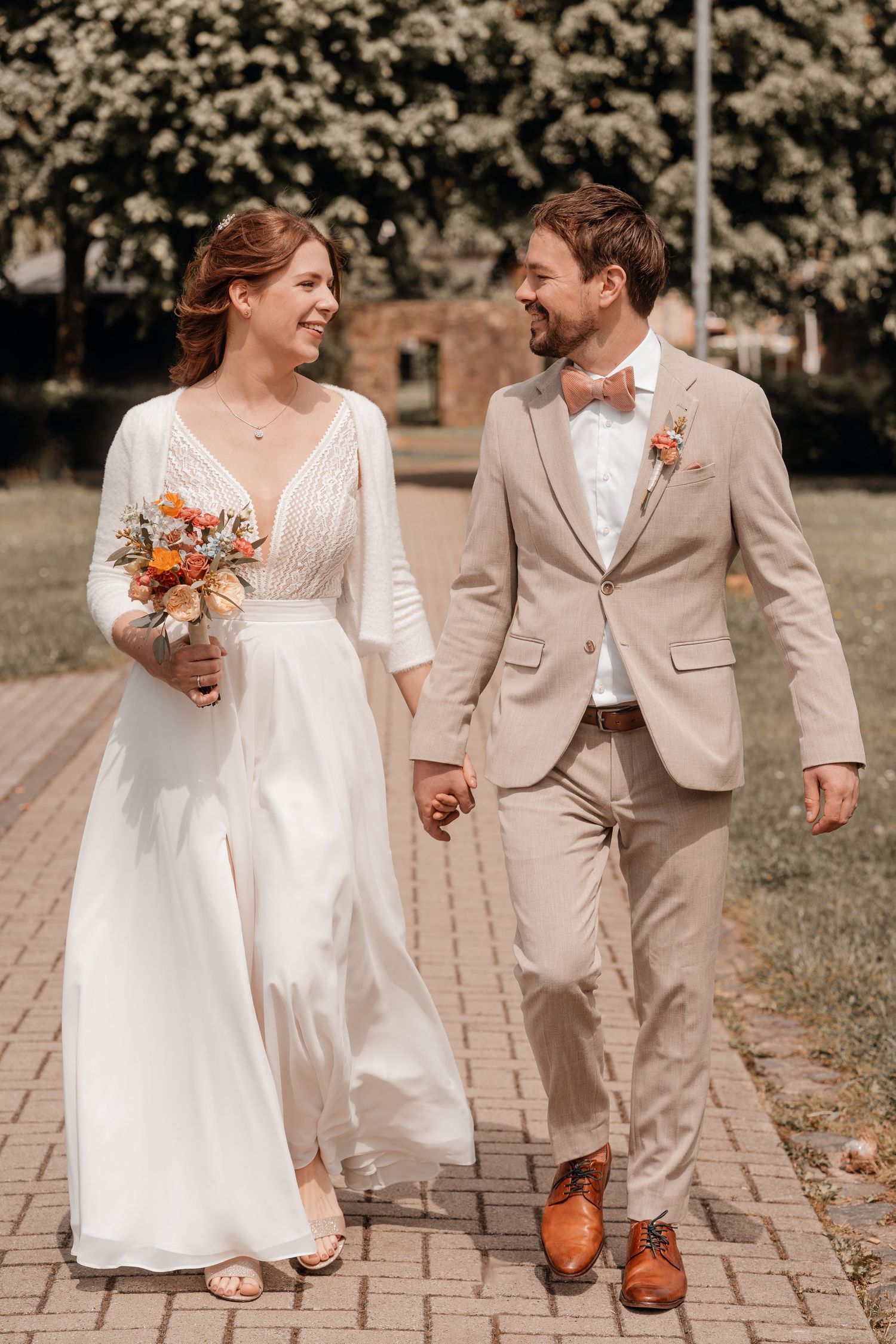 A bride and groom in white and beige attire walk hand in hand on a stone pathway while sharing joyful moments together.