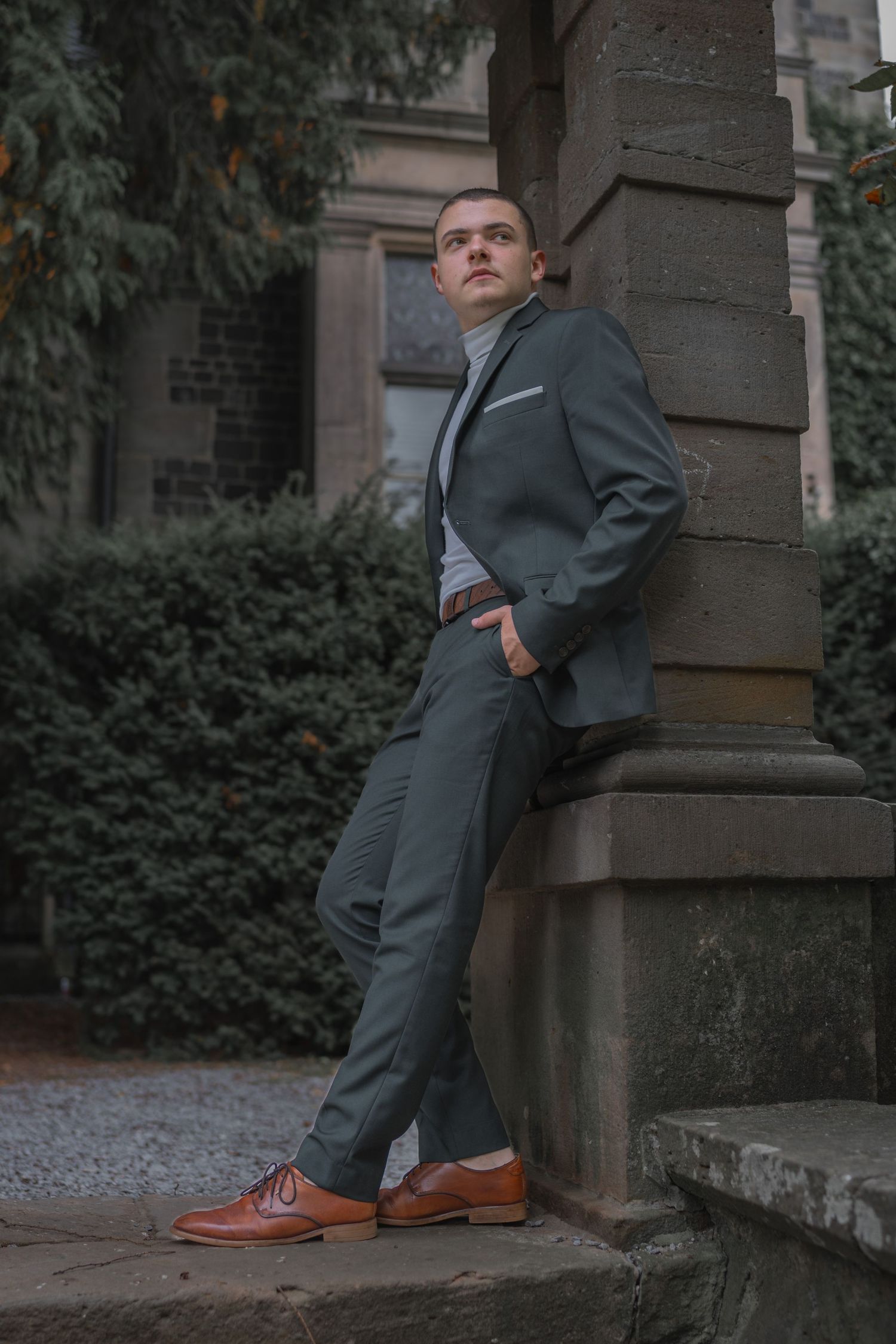 Fashion portrait of someone in a grey suit leaning against a stone pillar.