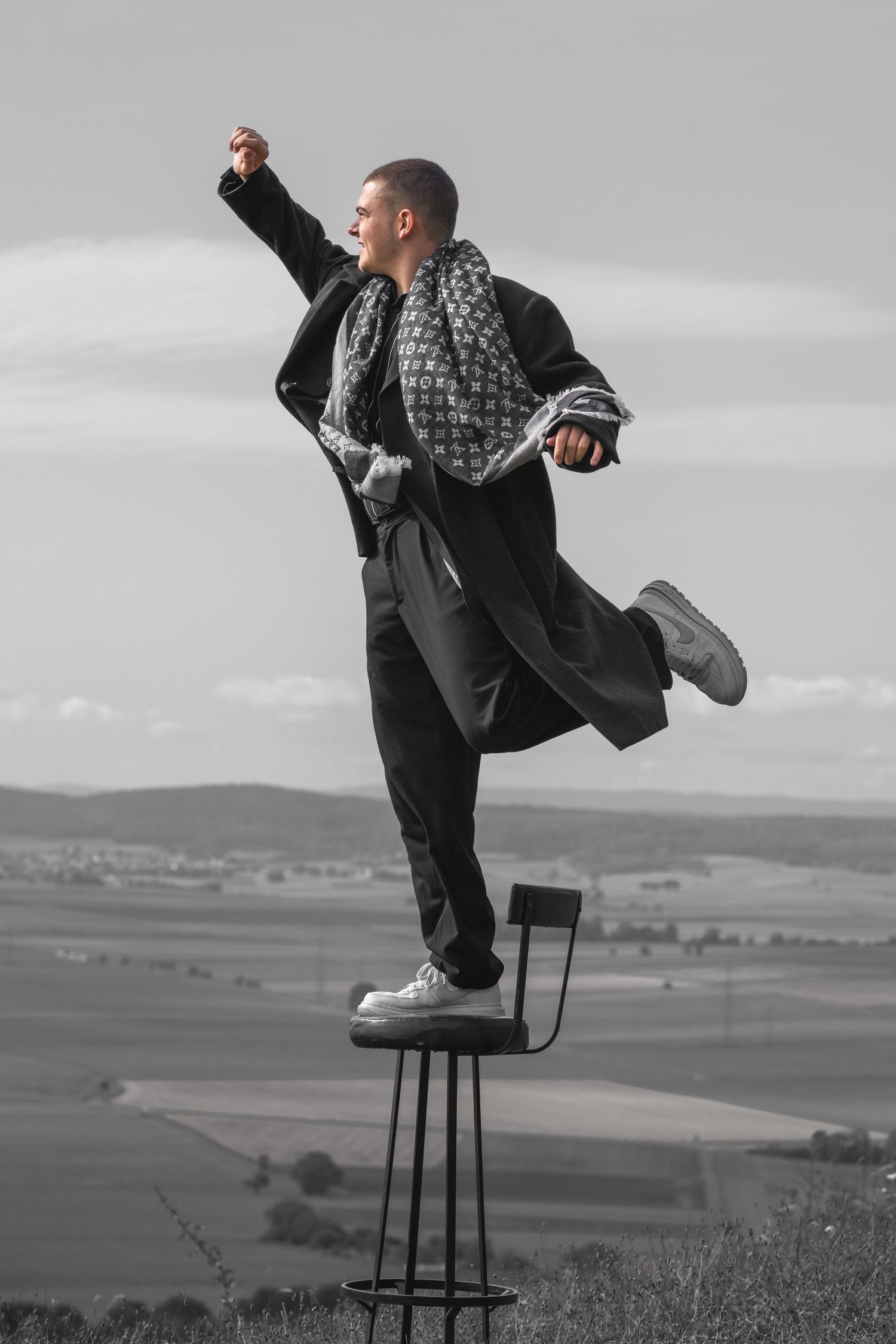 Artistic black and white photo of a person balancing dramatically on a tall stool in an outdoor setting.