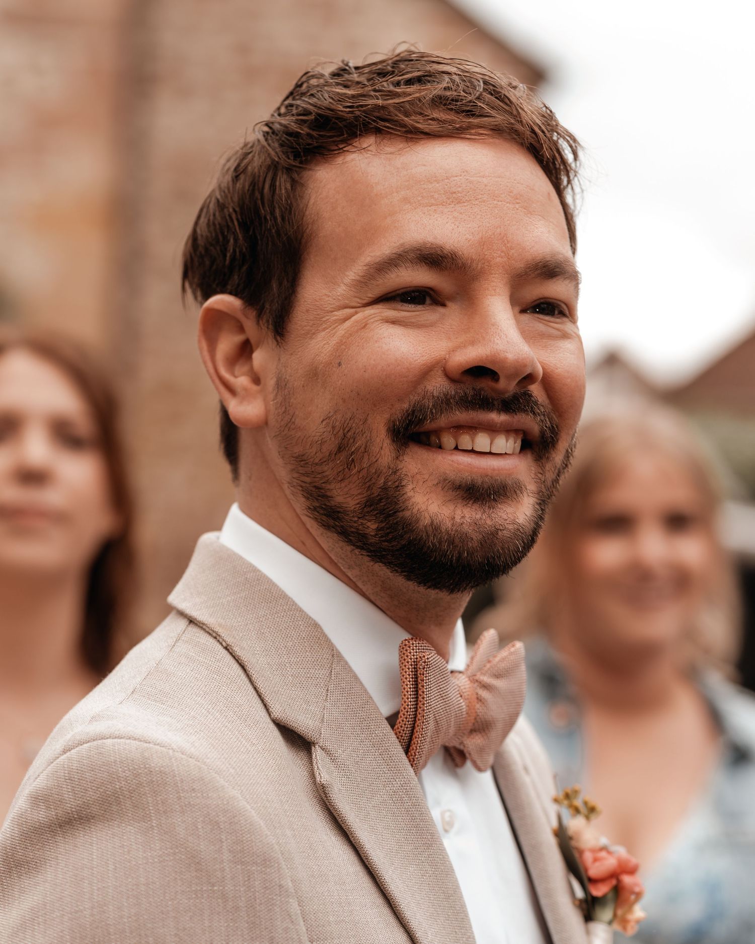 Groom in light beige suit smiling during wedding ceremony.