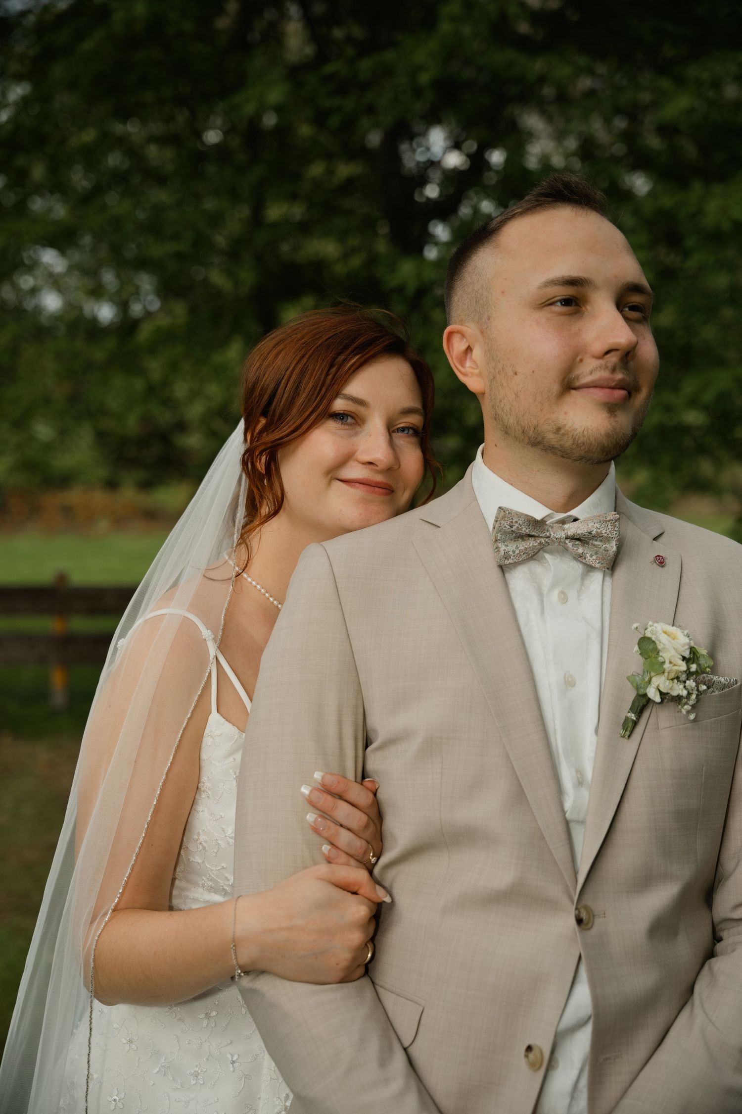 A bride in a white gown and veil leans on a groom in a beige suit with a gray bow tie outdoors.