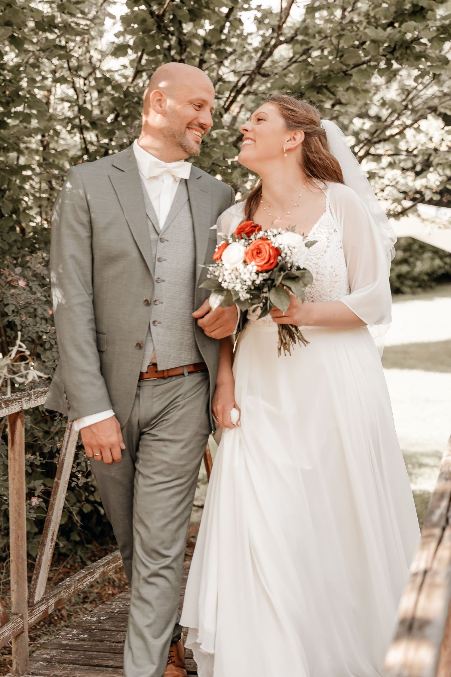 Happy bride and groom walking together outdoors, bride holding red and white bouquet, groom in gray suit with bow tie.