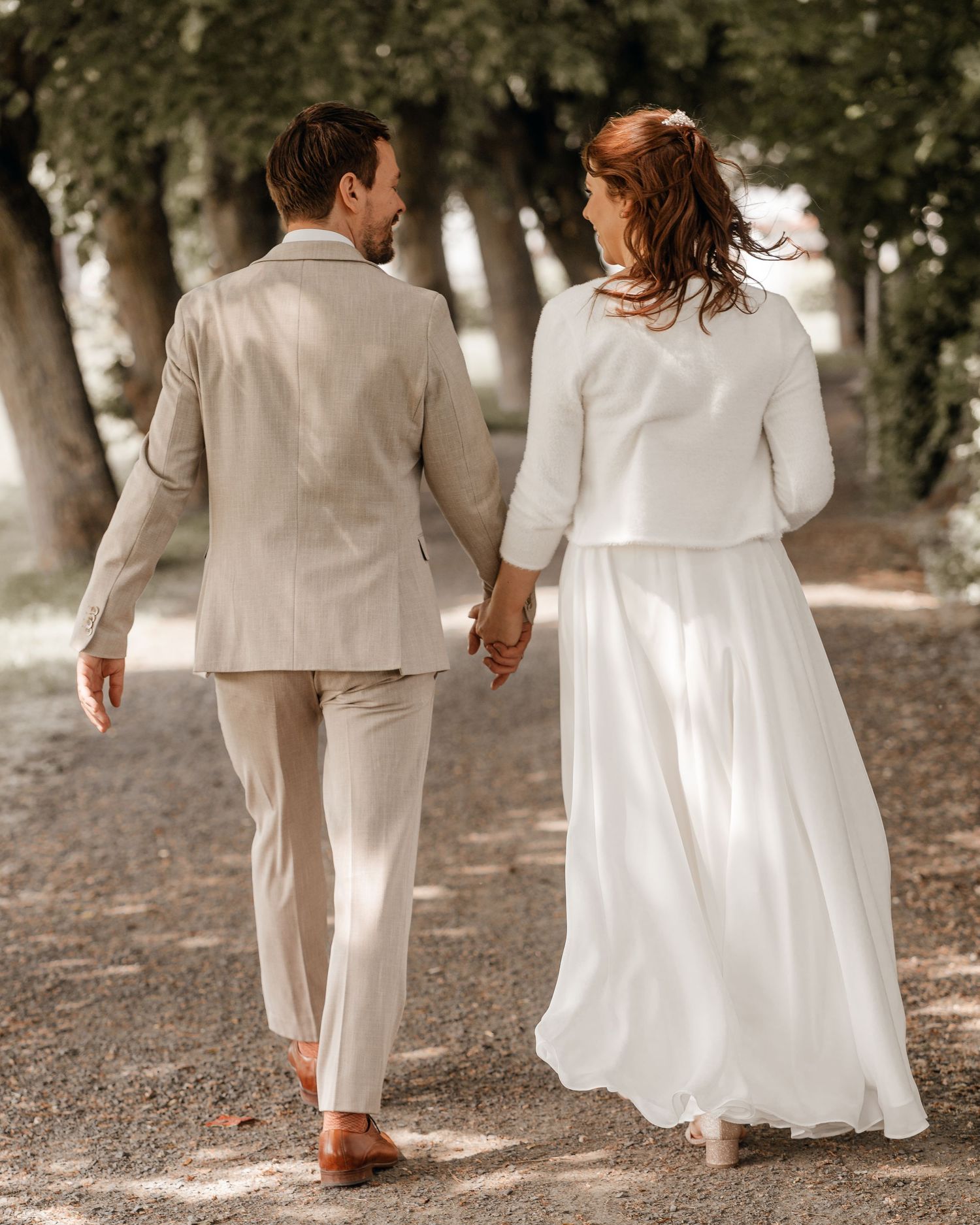 Couple in white wedding attire walking through a tree-lined path holding hands.