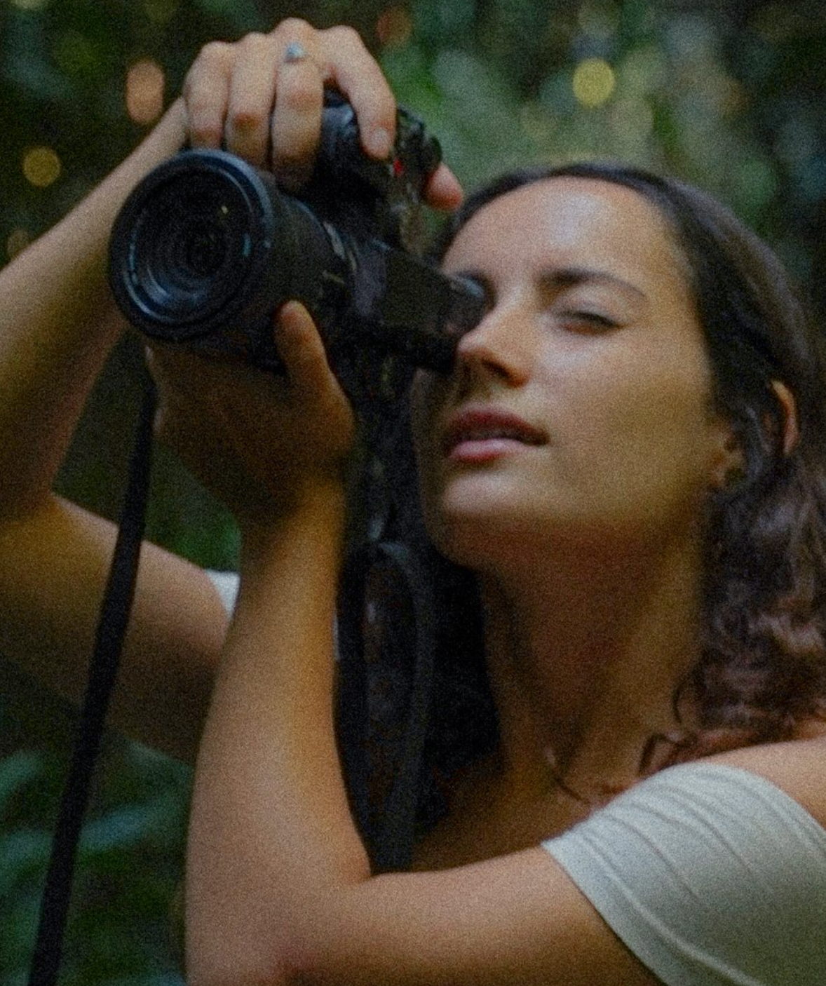 A photographer holds a camera lens against a blurred bokeh forest background in soft golden lighting.