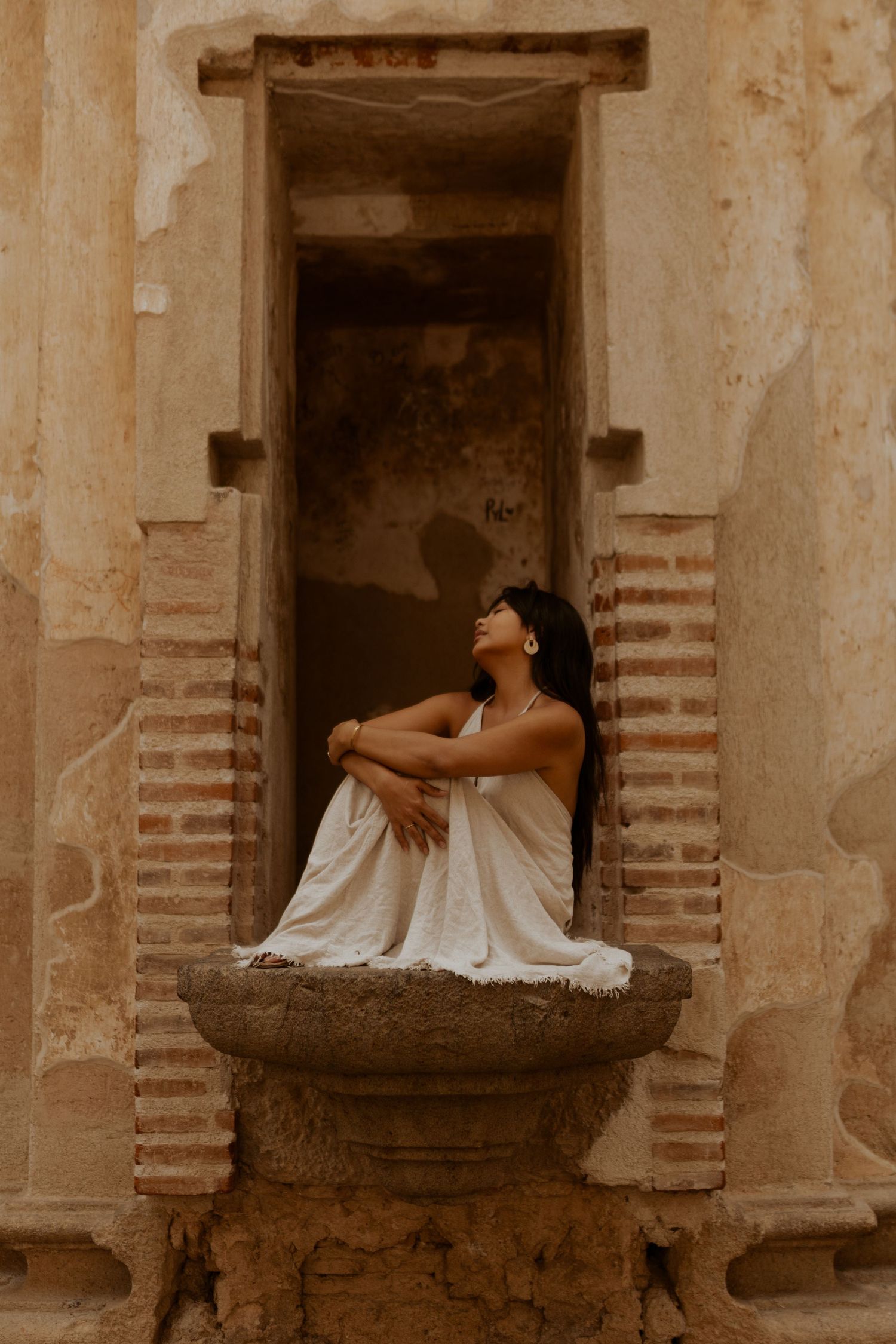 A figure draped in white fabric sits contemplatively on an old stone balcony against a rustic textured wall.