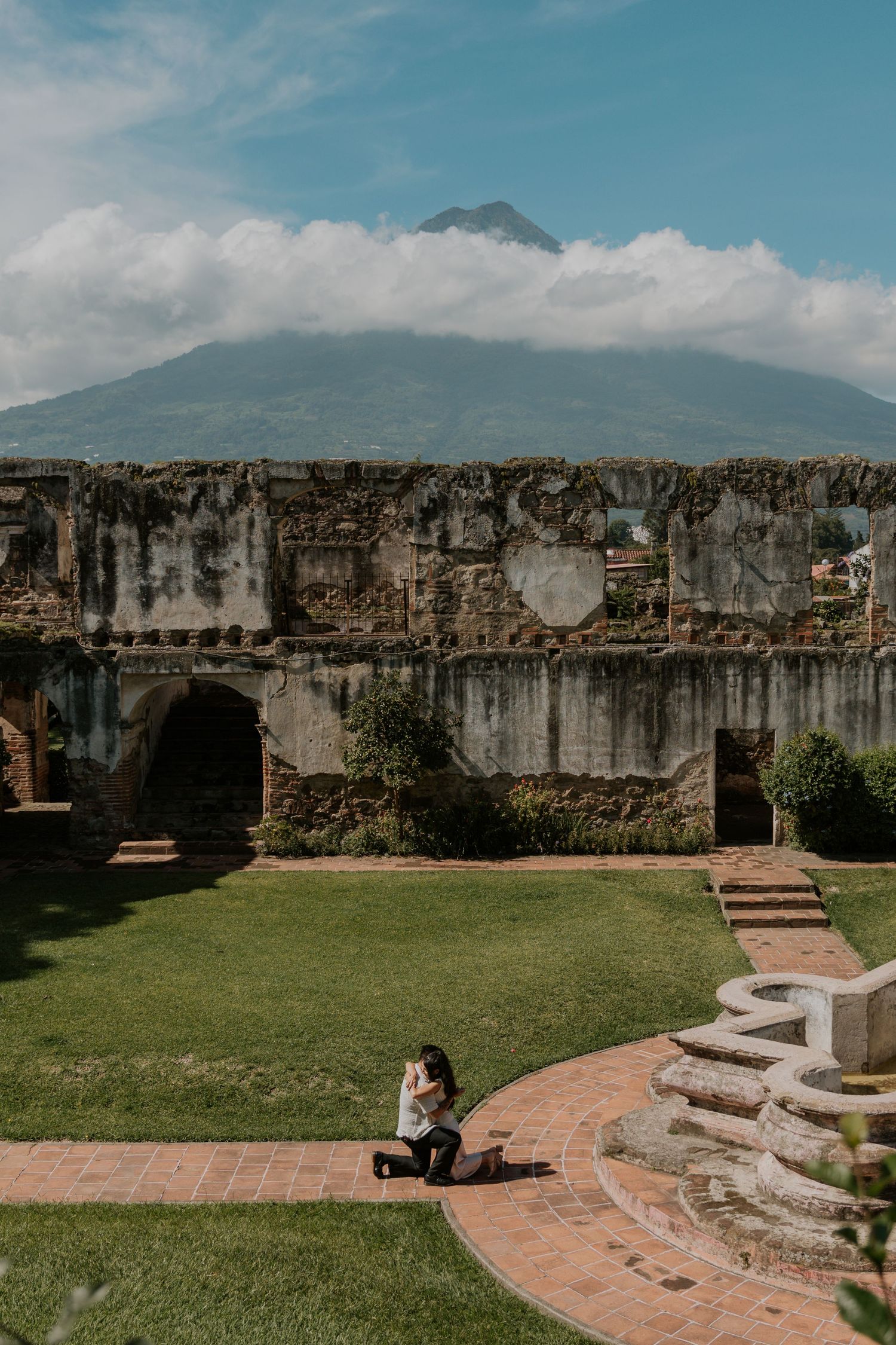 Person sitting in ruins courtyard with lush lawn, stone arches, and volcano peak emerging through clouds in background.
