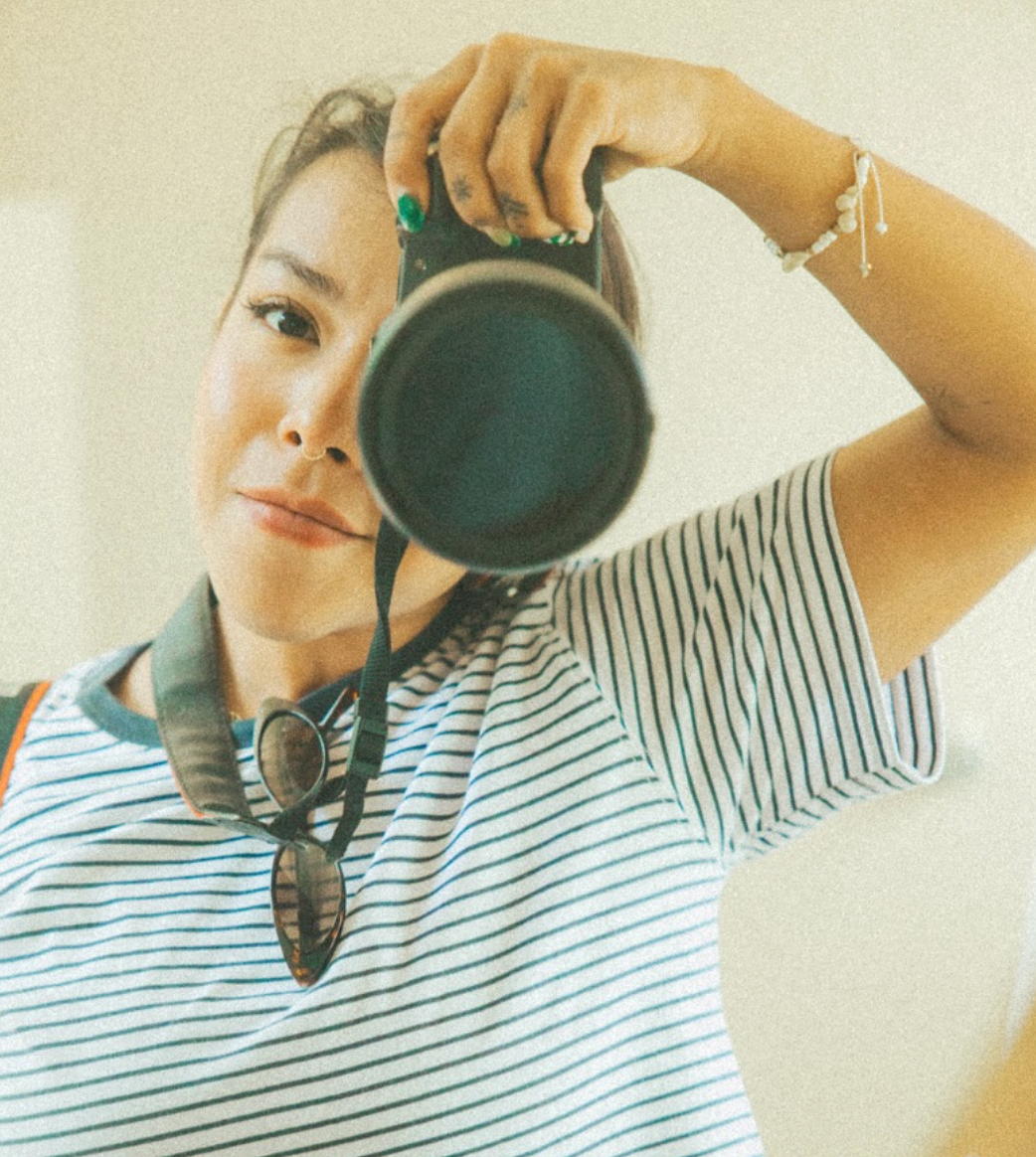 Woman in striped shirt holds DSLR camera up to her face, wearing sunglasses around neck, taking a mirror selfie.