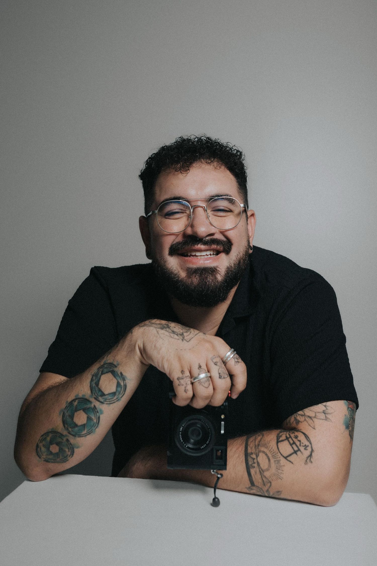Portrait against gray wall shows someone wearing glasses and black t-shirt with visible arm tattoos, smiling while leaning on table.