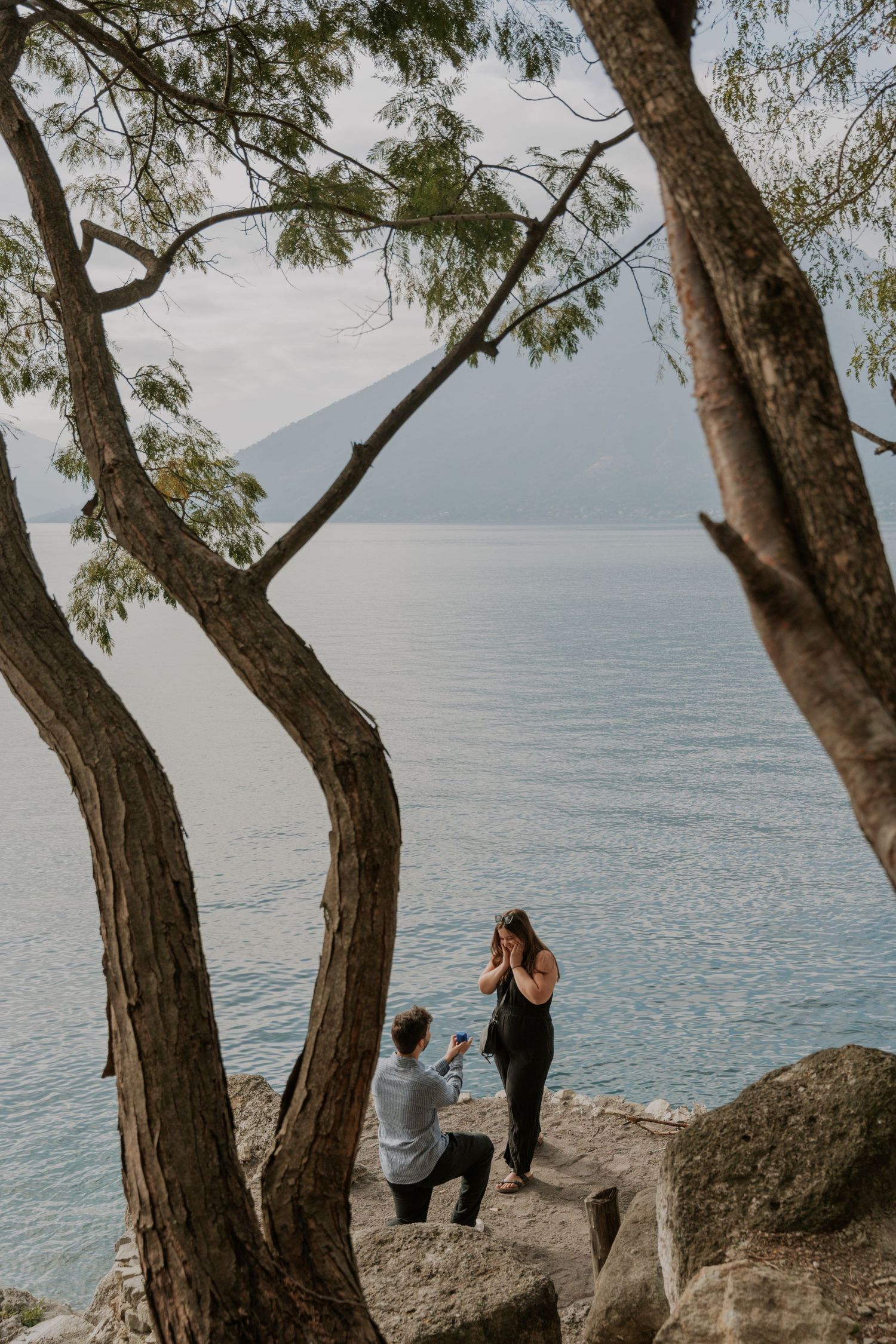 A couple shares a romantic moment on rocky cliffs overlooking a serene lake with curved trees framing the scene.