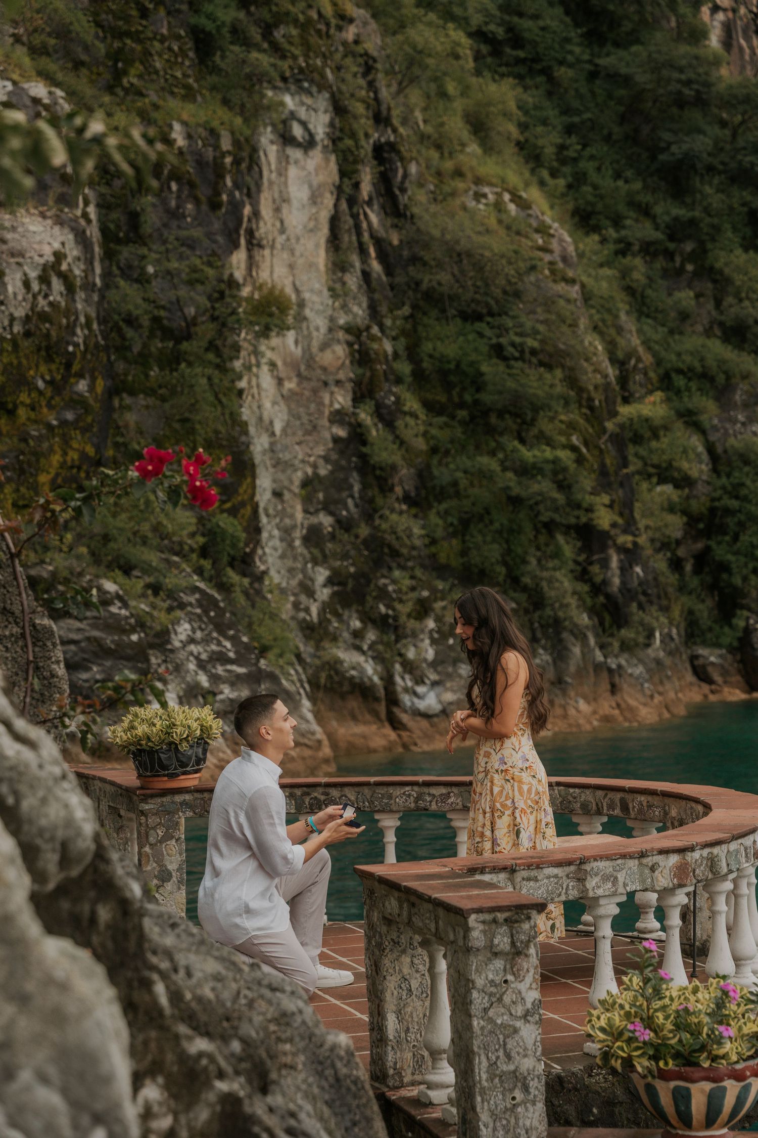 A couple shares a romantic moment on a terrace overlooking the Amalfi Coast with stone columns and vibrant flowers.