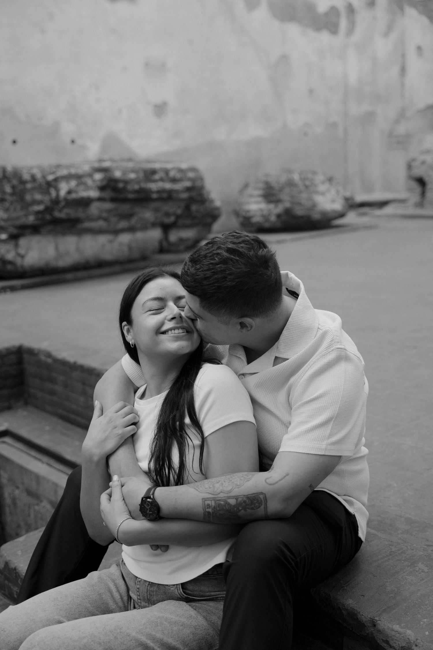 Black and white photo of a couple embracing, man kissing woman's cheek as she smiles with eyes closed outdoors.