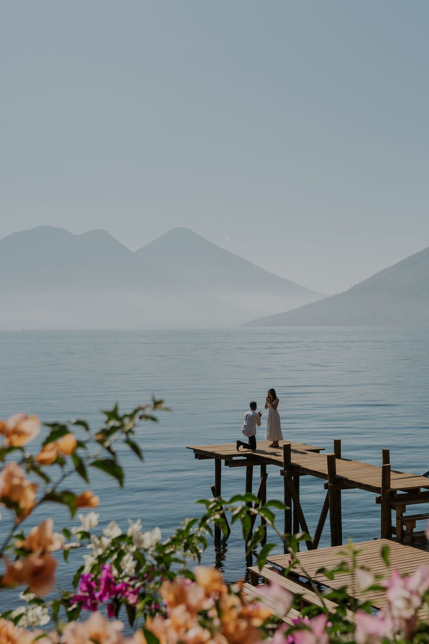 Two silhouettes stand on a wooden pier overlooking a misty lake surrounded by mountains and colorful flowers in foreground.