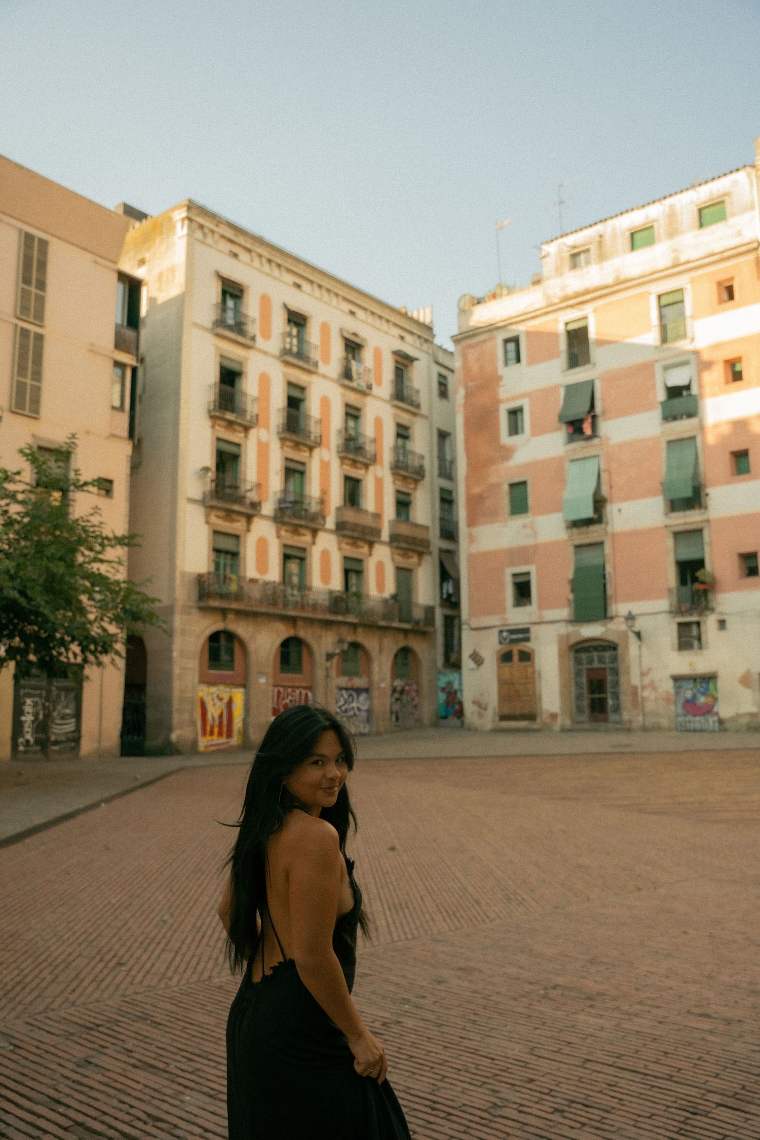 Young woman in black backless dress poses in a sunlit European cobblestone plaza surrounded by colorful aged buildings.