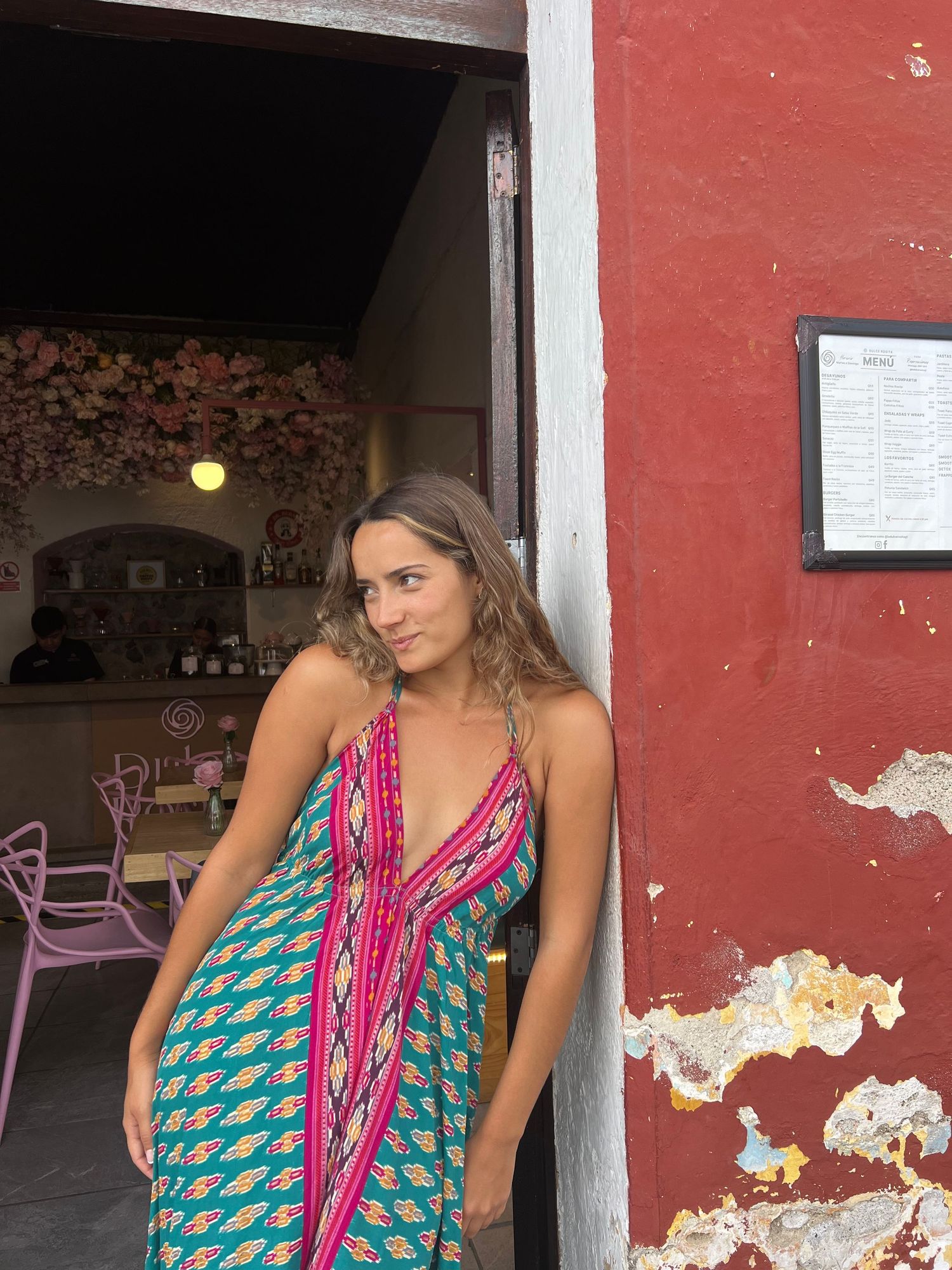 A person wearing a colorful geometric print maxi dress poses in a doorway with pink and teal patterns against a red wall.