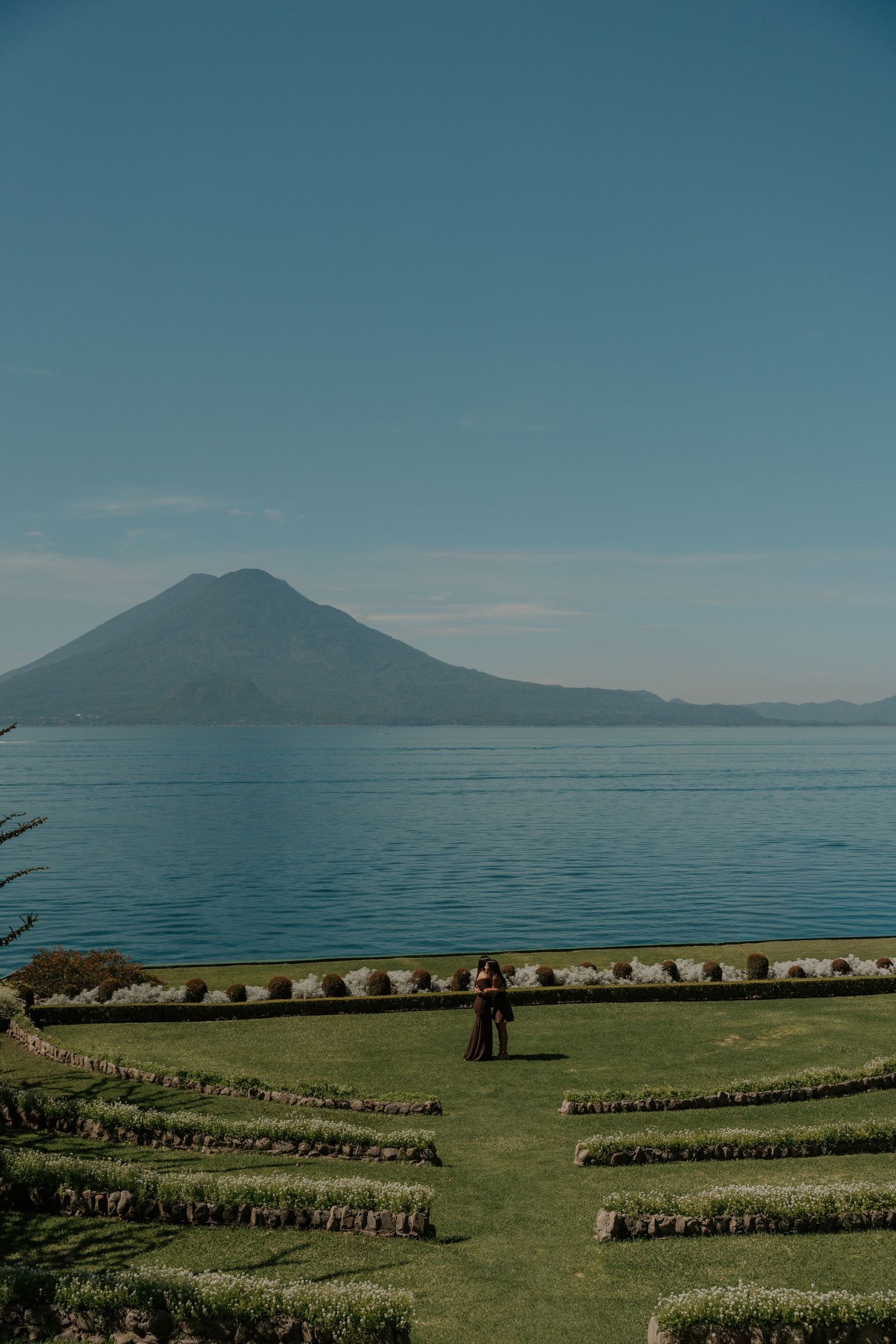 Panoramic view of sheep grazing on green pastures near a tranquil lake with mountains in the distance.